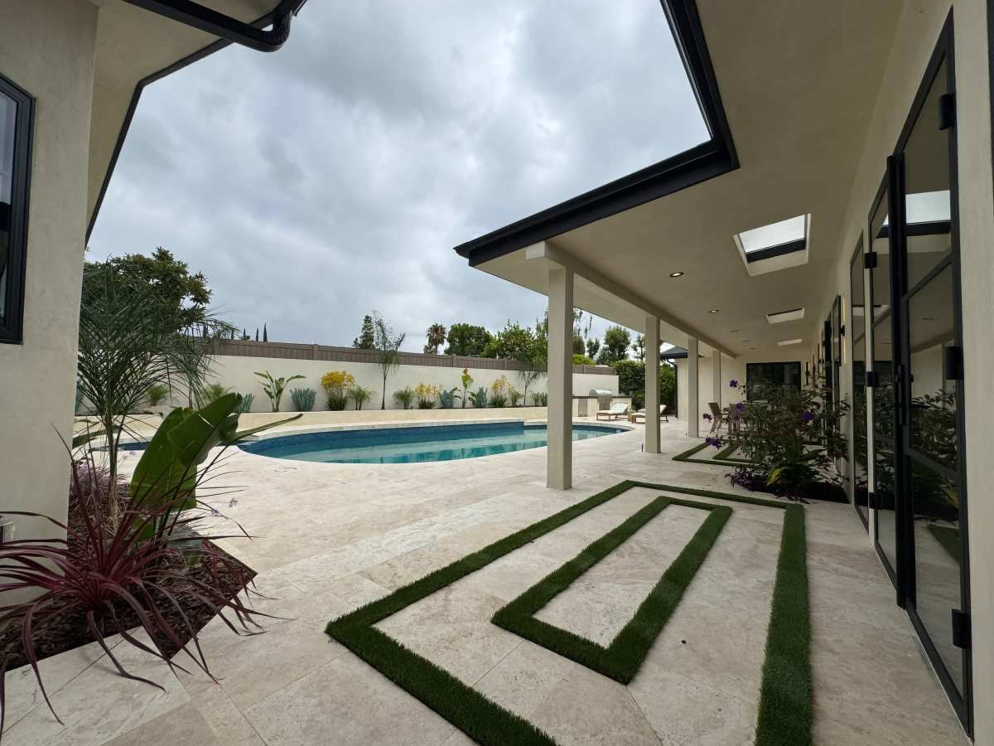 The image shows a backyard with a swimming pool surrounded by landscaped greenery and a patio area featuring stone flooring and decorative grass strips.
