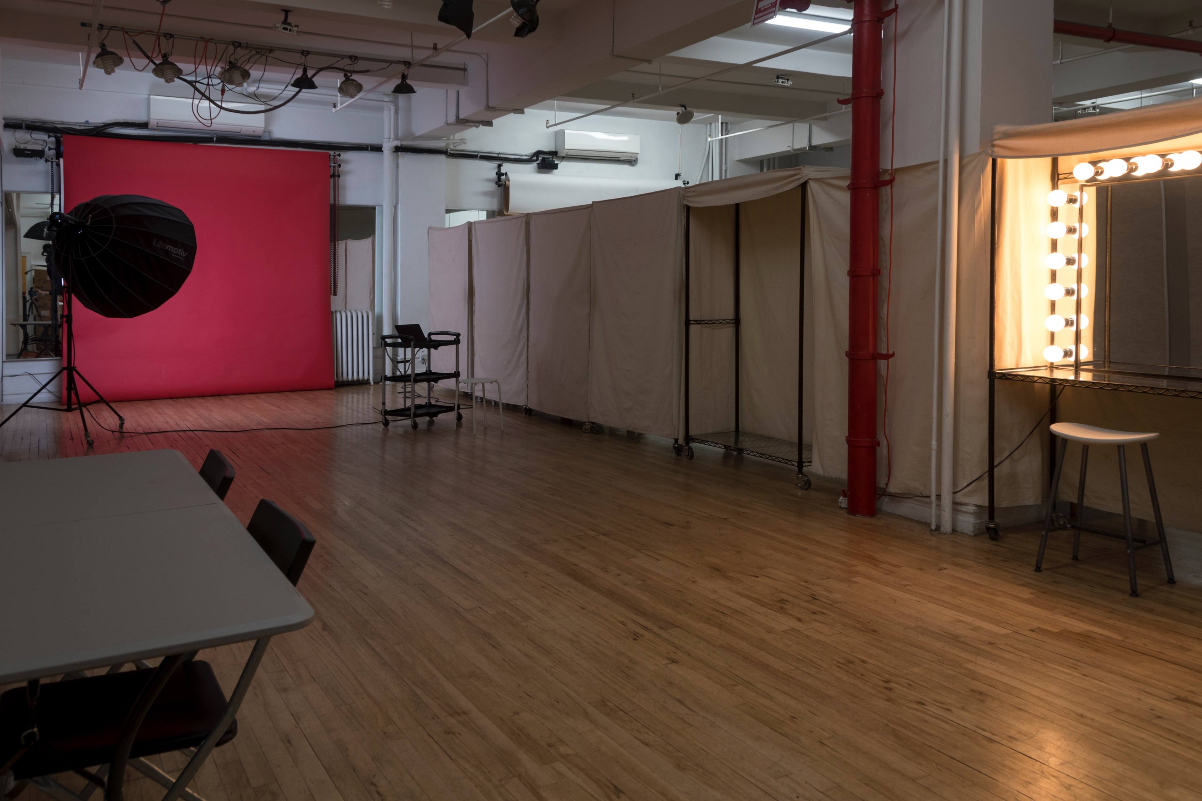 The image shows a photography studio with a red backdrop, a light setup, and a dressing area with illuminated mirrors.