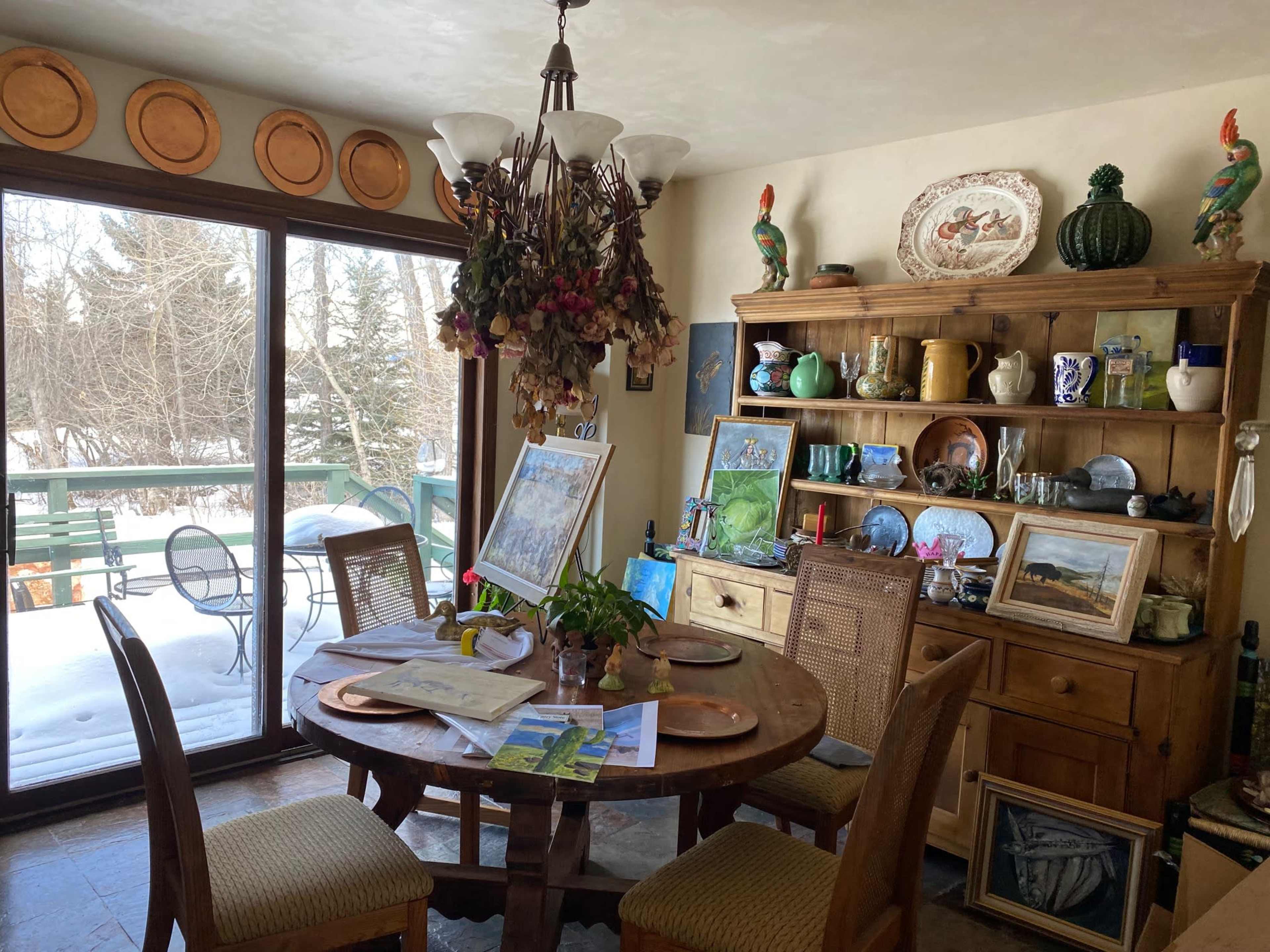 A dining area features a round wooden table surrounded by chairs, with a view of a snowy outdoor scene through large windows and various decorative items displayed on shelves.