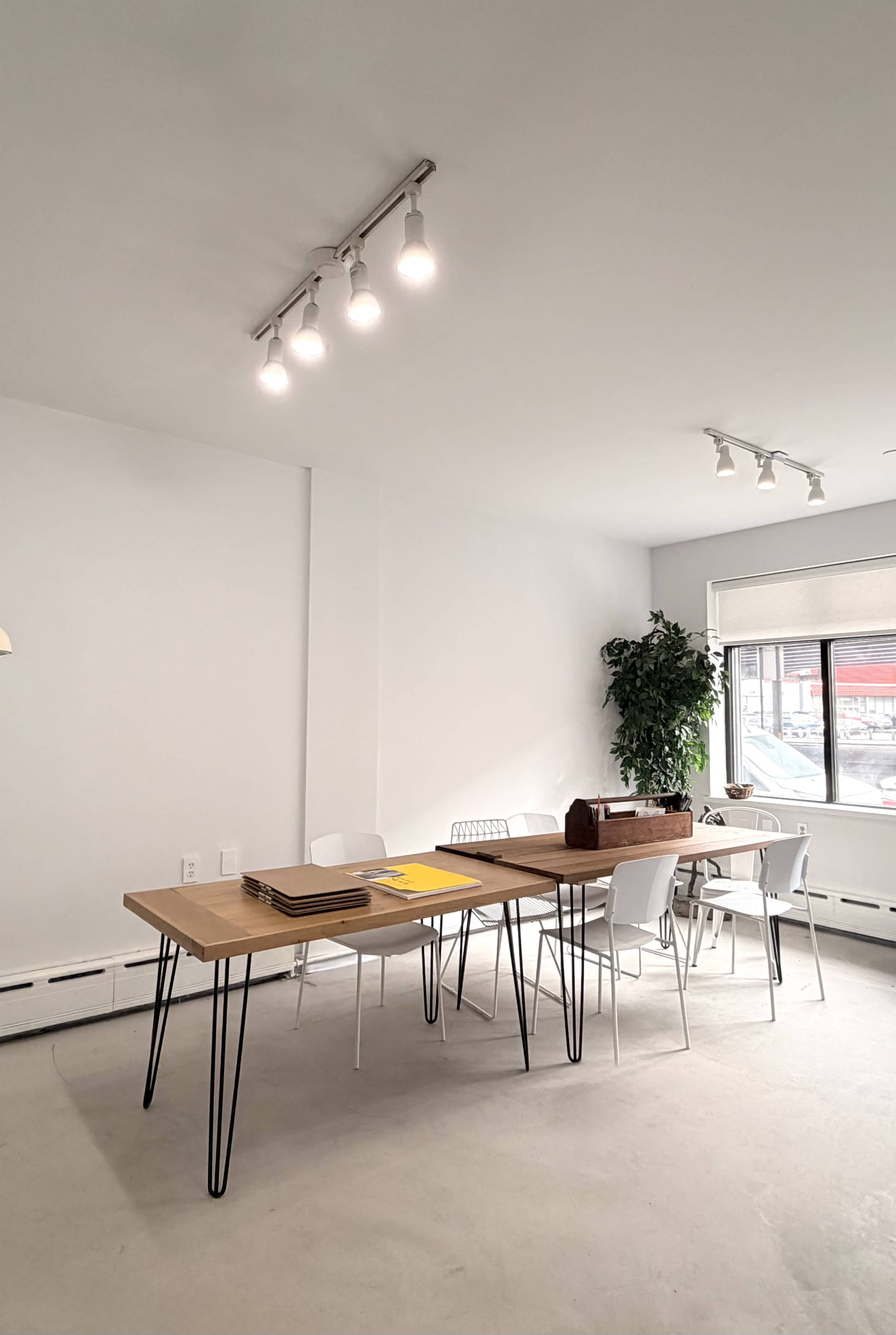 A spacious room features a wooden table with modern white chairs, accompanied by a window and a large potted plant.
