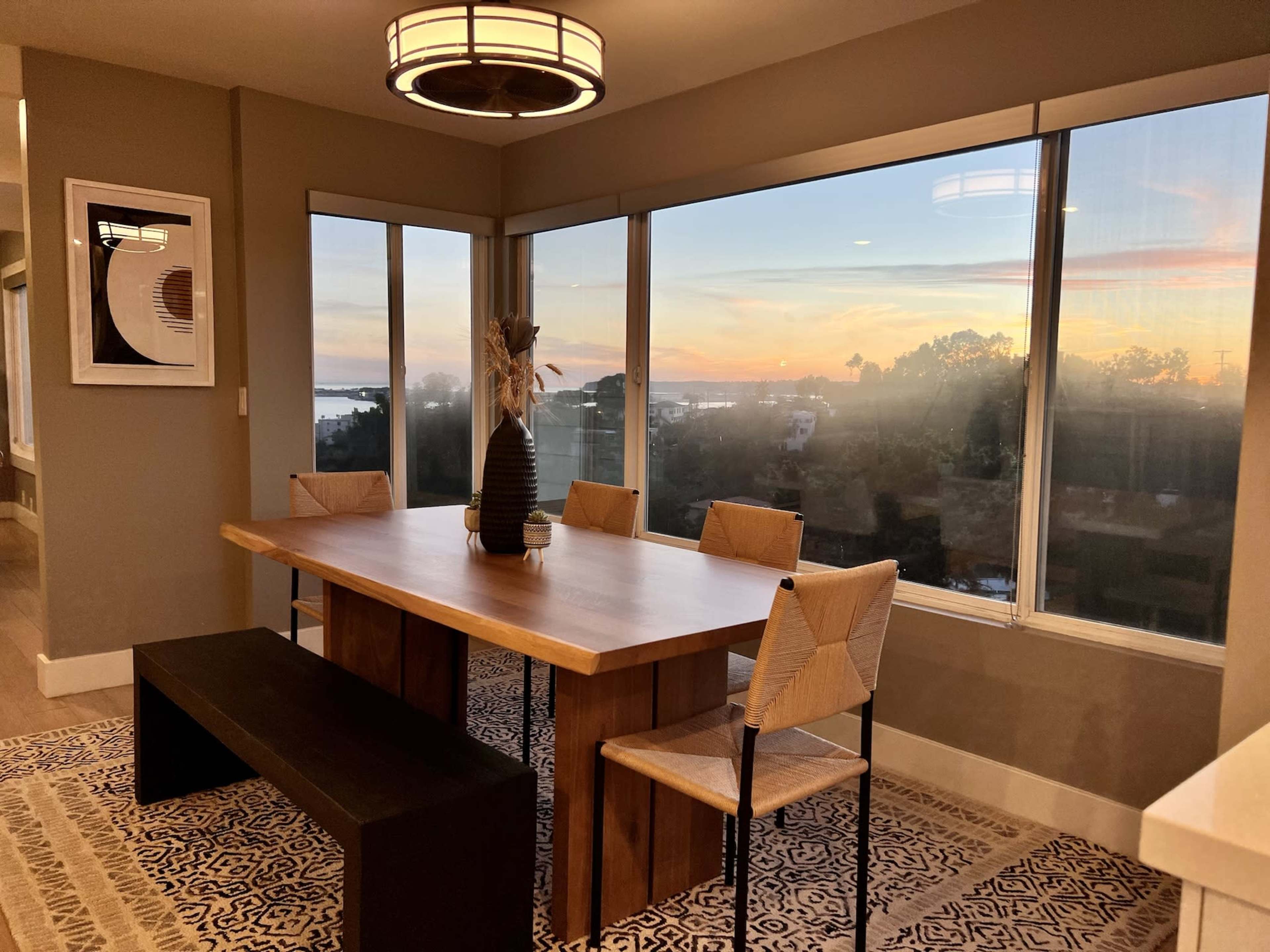 A wooden dining table with woven chairs is positioned next to large windows overlooking a sunset-lit landscape.