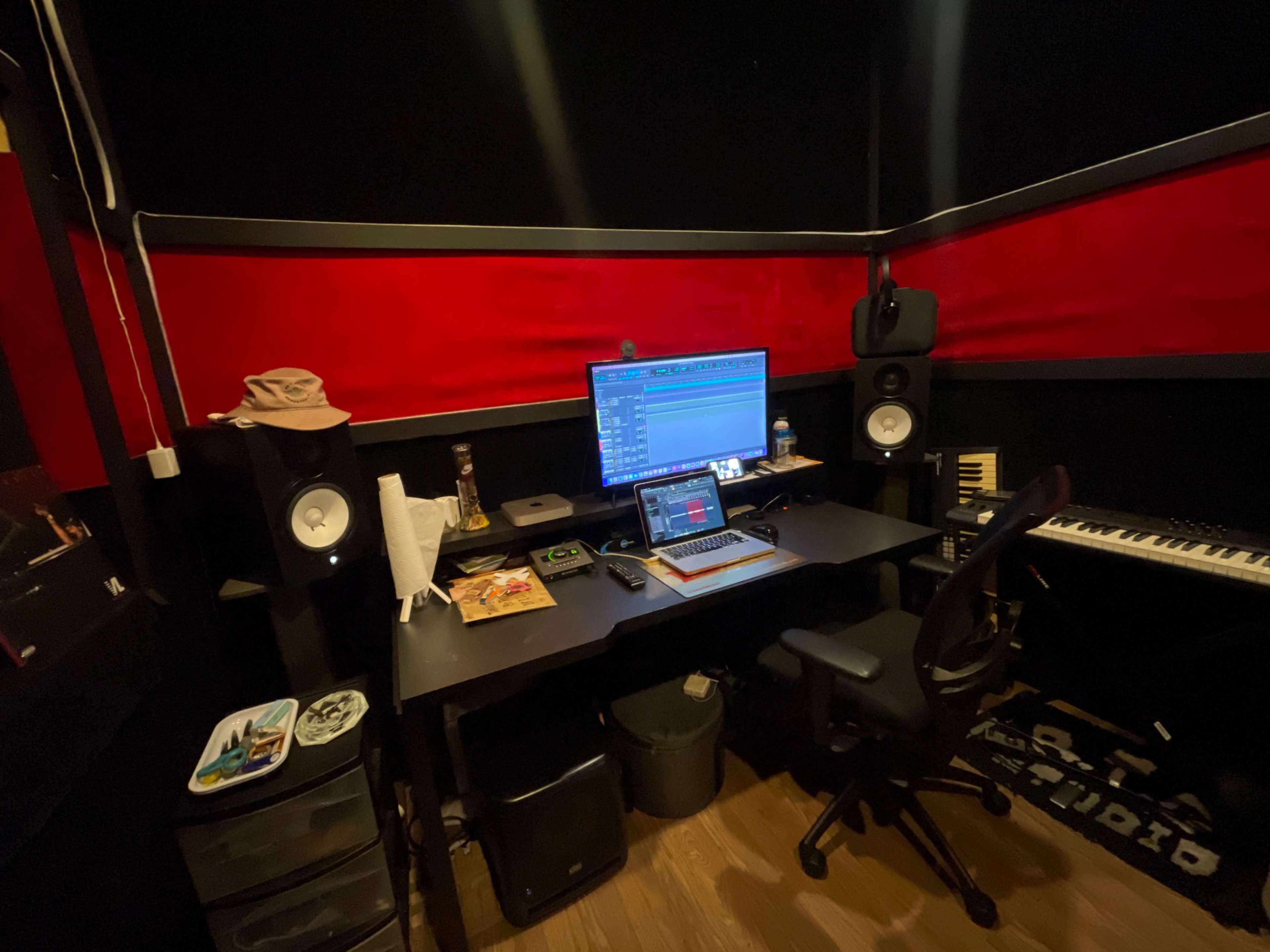 The image shows a music production studio featuring a desk with a computer monitor, speakers, a keyboard, and various music production equipment against a backdrop of red and black walls.