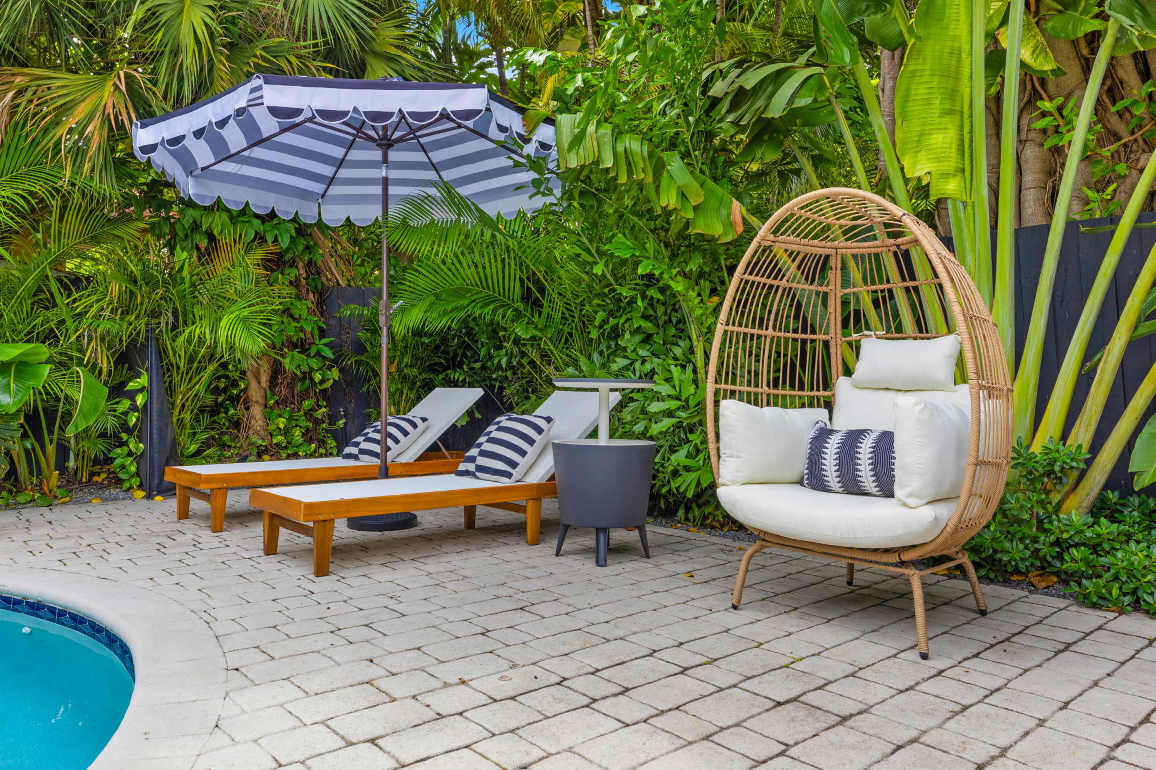 The scene features a poolside area with two striped lounge chairs, a round table, a large striped umbrella, and a unique, egg-shaped chair surrounded by lush greenery.