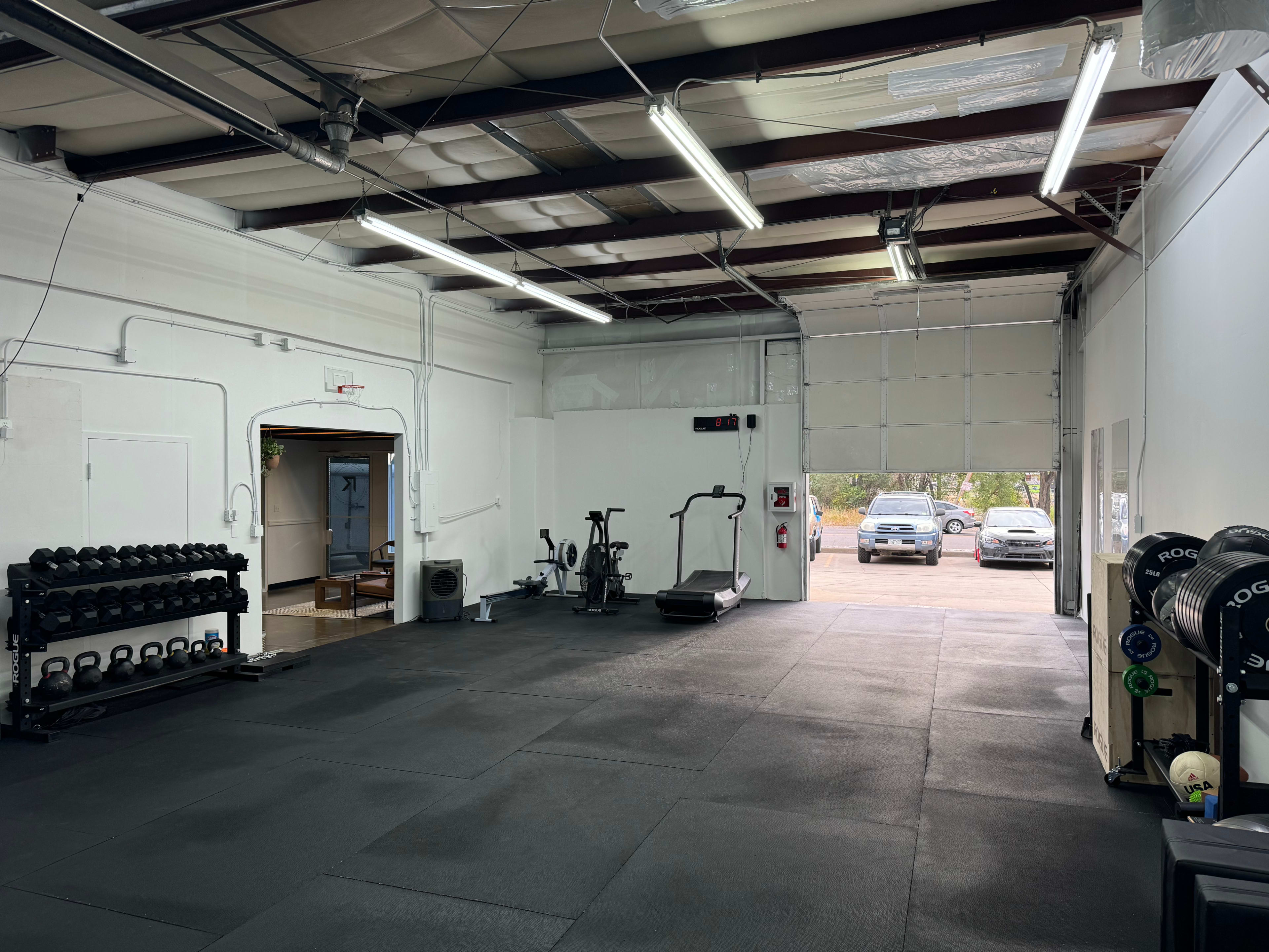 The image shows an empty gym space with exercise equipment, including dumbbells, a treadmill, and an elliptical, set against a white wall.