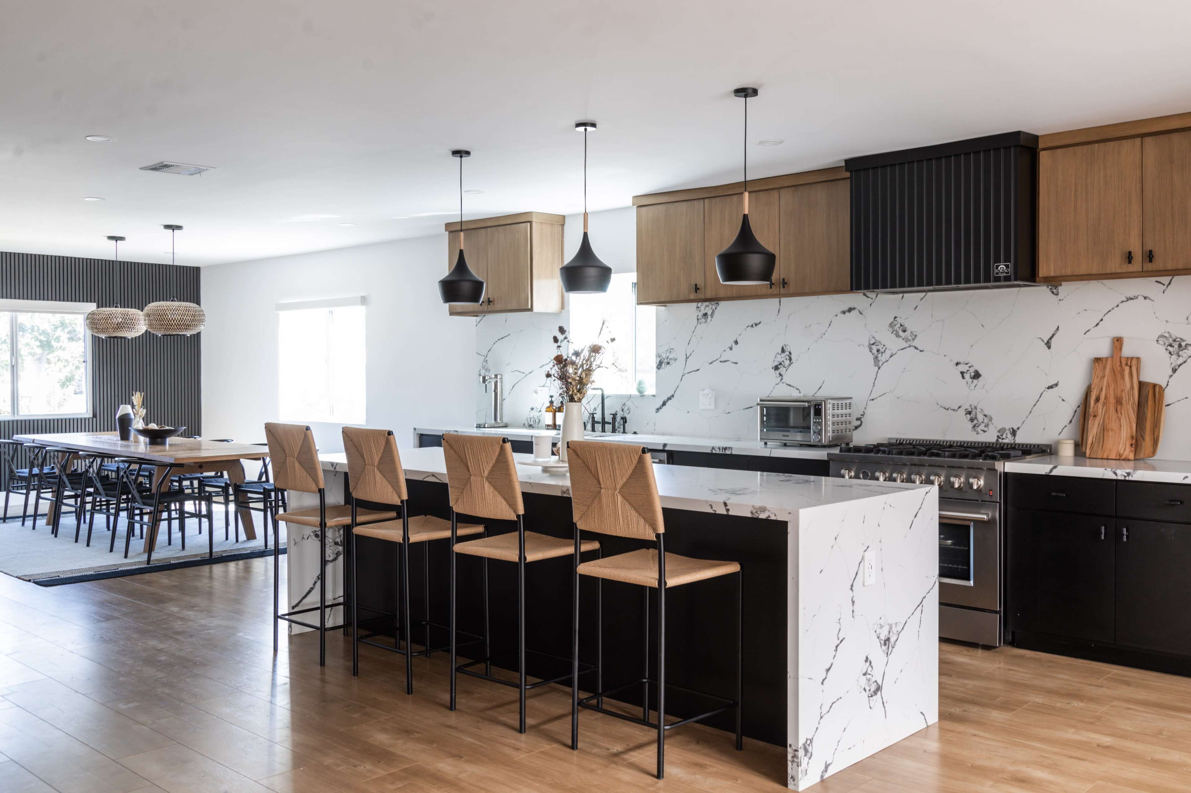 A modern kitchen features a large island with black bar stools, marble countertops, and wood cabinetry, adjoining a dining area with a long table.