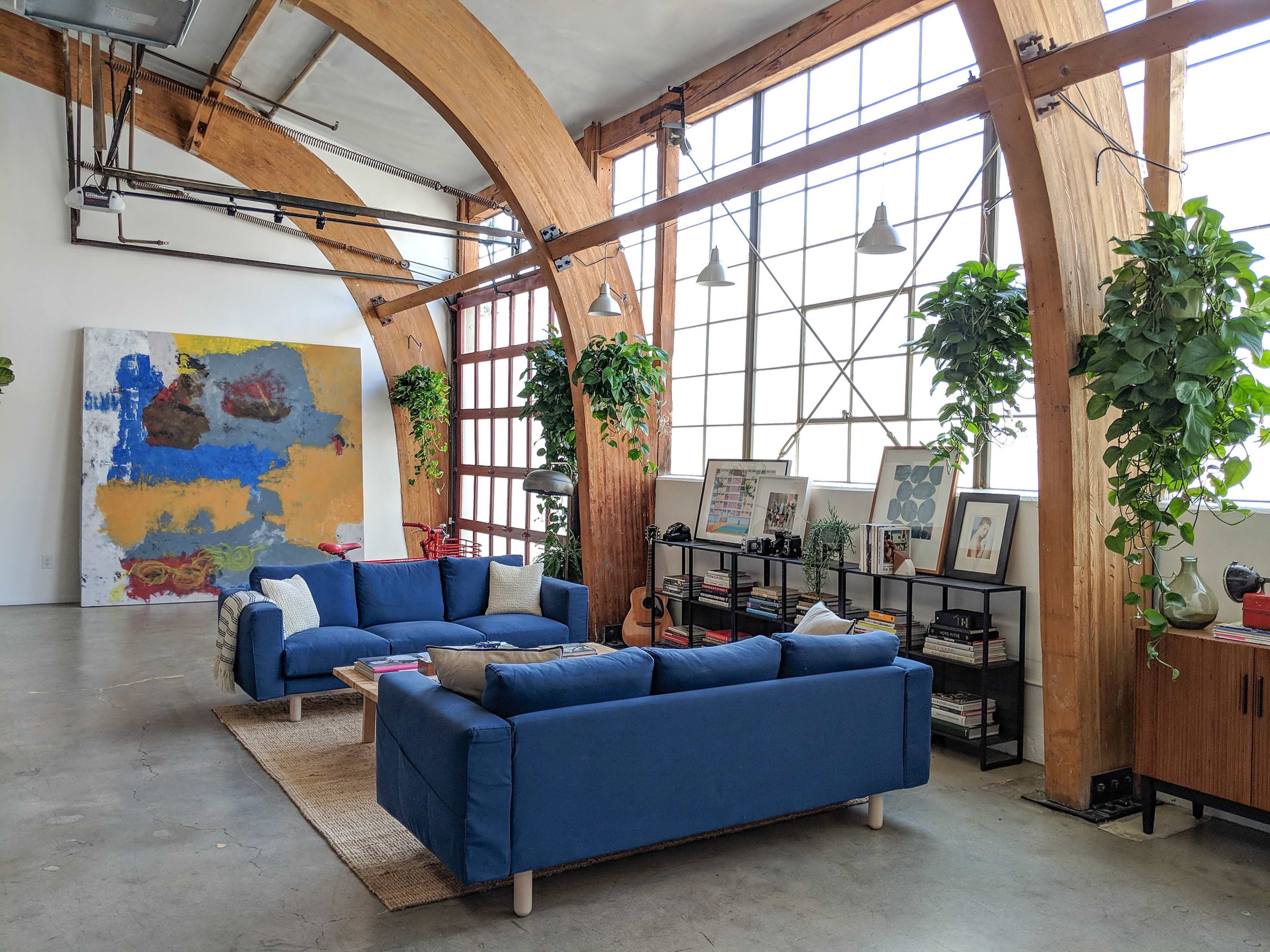 A spacious living room with large windows, wooden arches, two blue sofas, and various plants, alongside artwork on the walls.