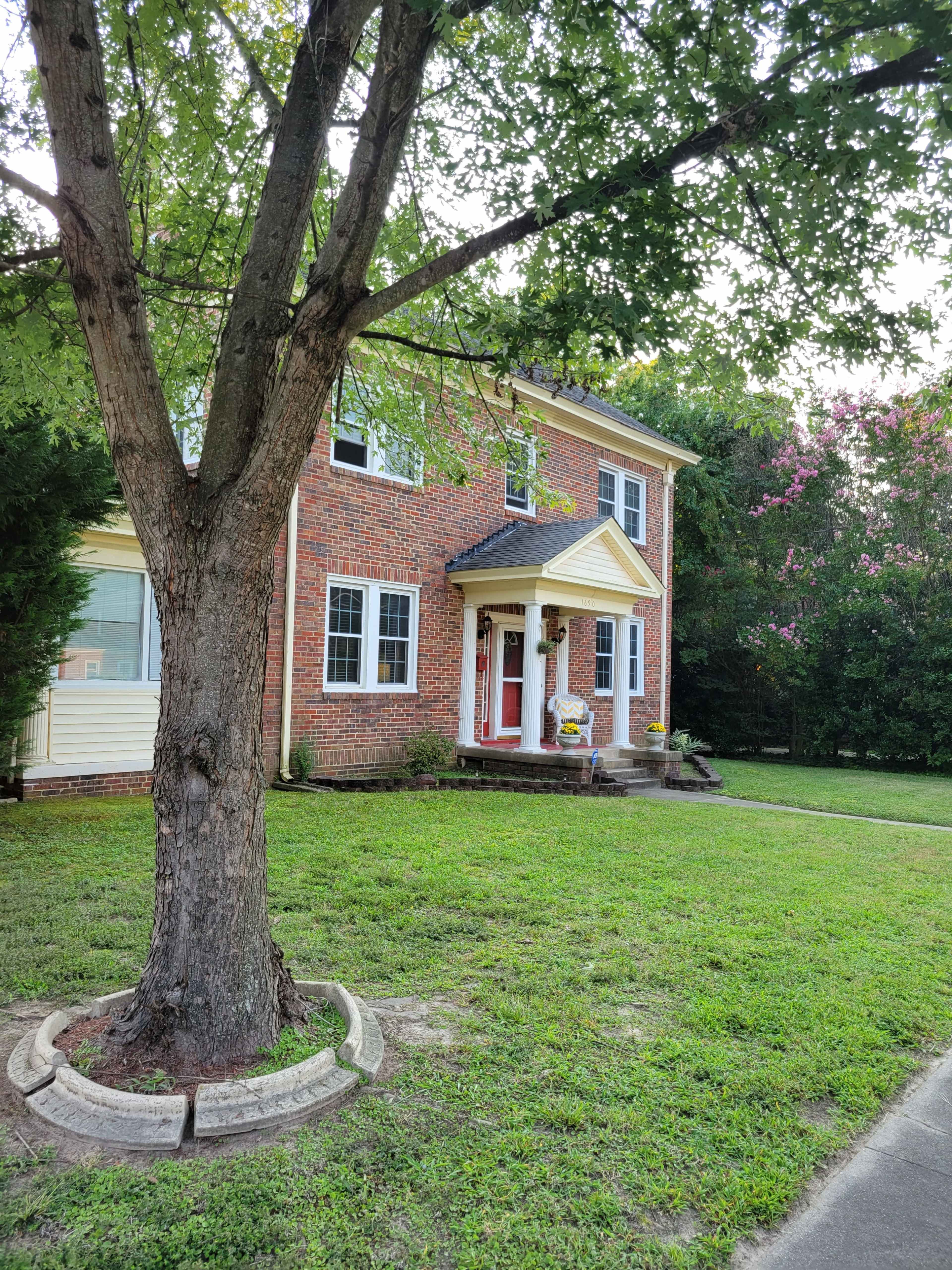A two-story brick house with a red front door is surrounded by green grass and trees.