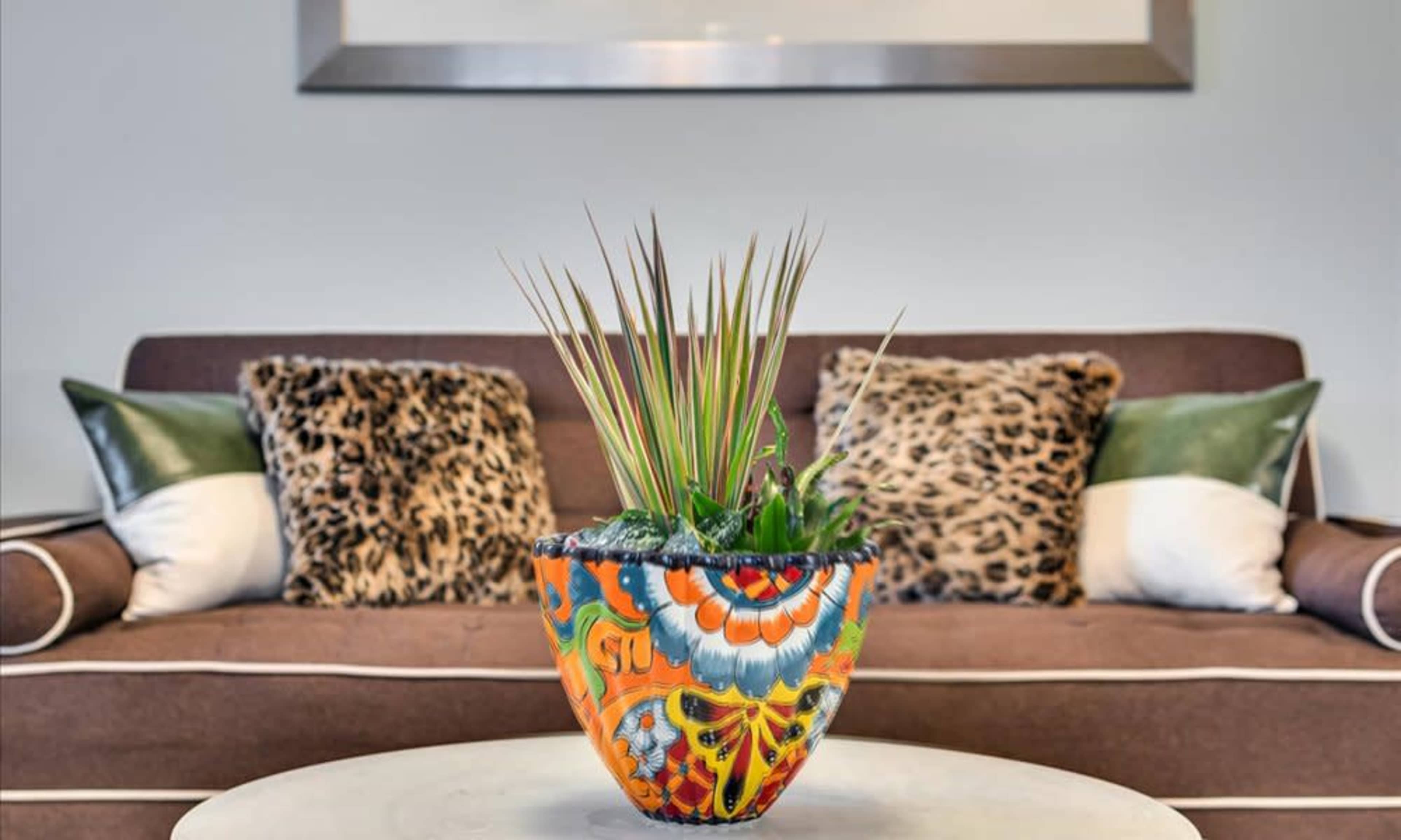 A colorful, patterned pot with plants sits on a coffee table in front of a brown sofa adorned with leopard print cushions.
