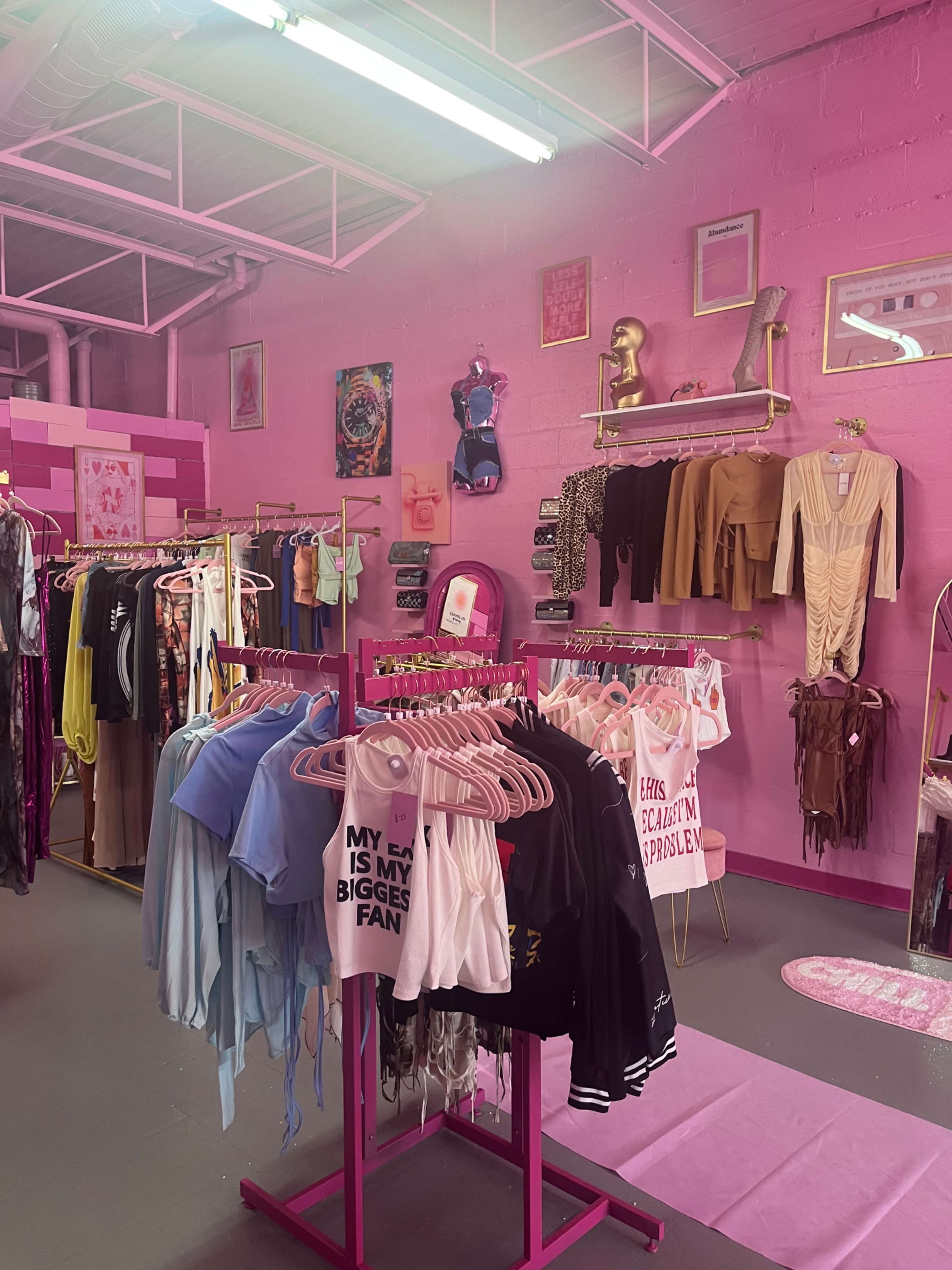 The image shows a clothing boutique with pink walls, featuring racks of colorful apparel and accessories.