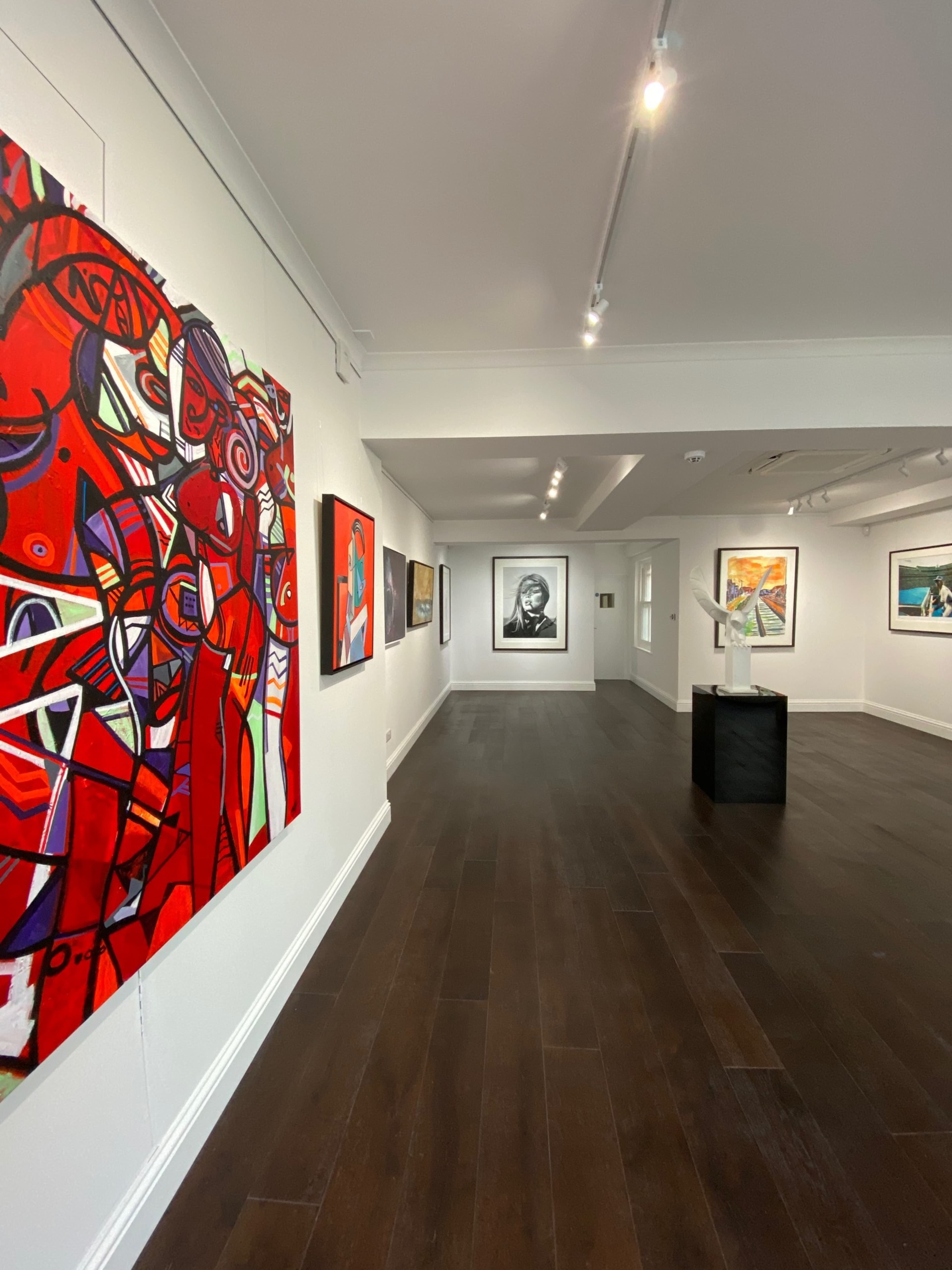 The image depicts an art gallery featuring various paintings on the walls and a sculpture in the center, with a wooden floor and ample lighting.
