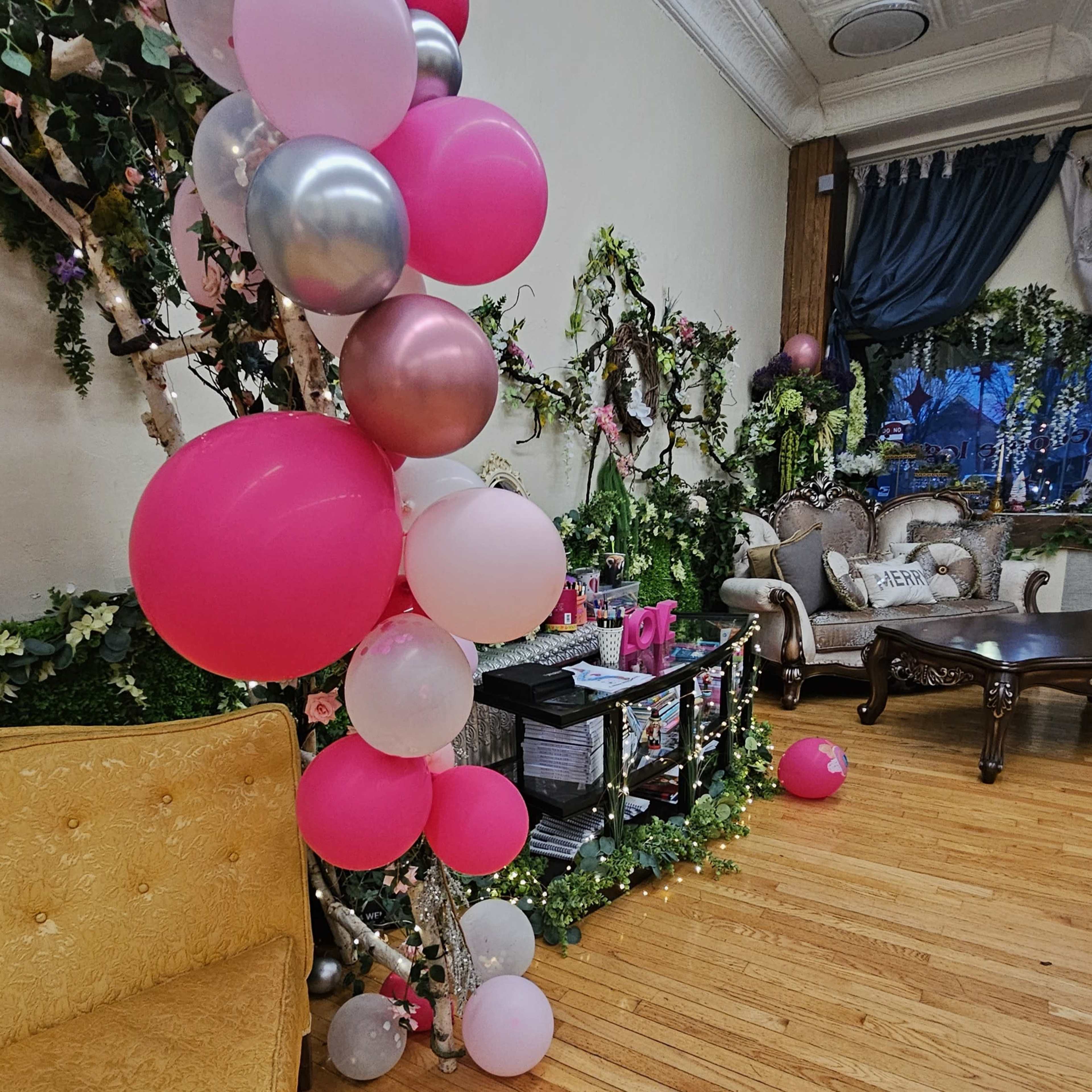 The image shows a decorated indoor space featuring a cluster of pink and silver balloons alongside a seating area with elegant furniture and floral arrangements.