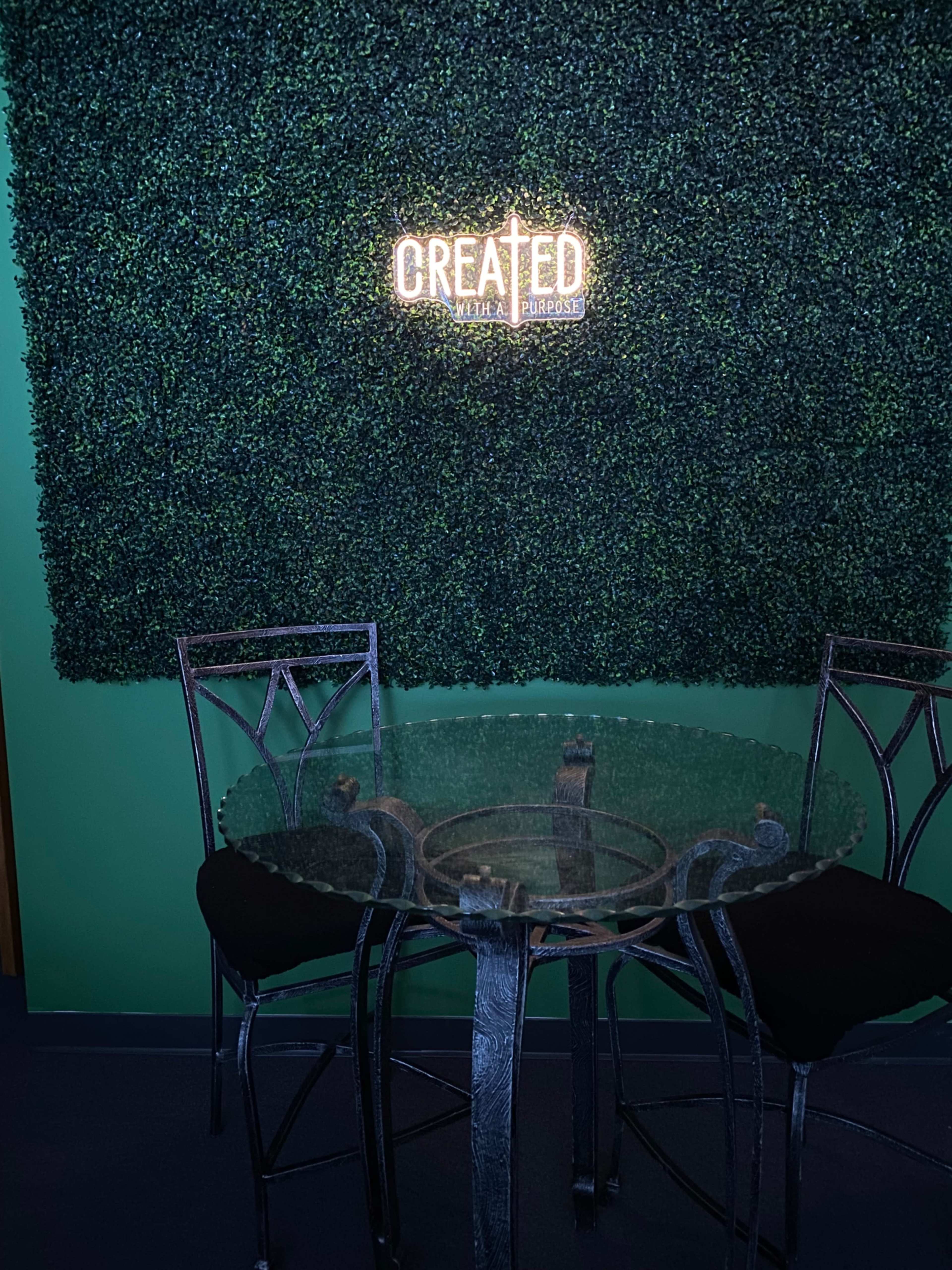 A round glass table with black chairs is positioned in front of a green wall featuring a lit sign that reads "CREATED."