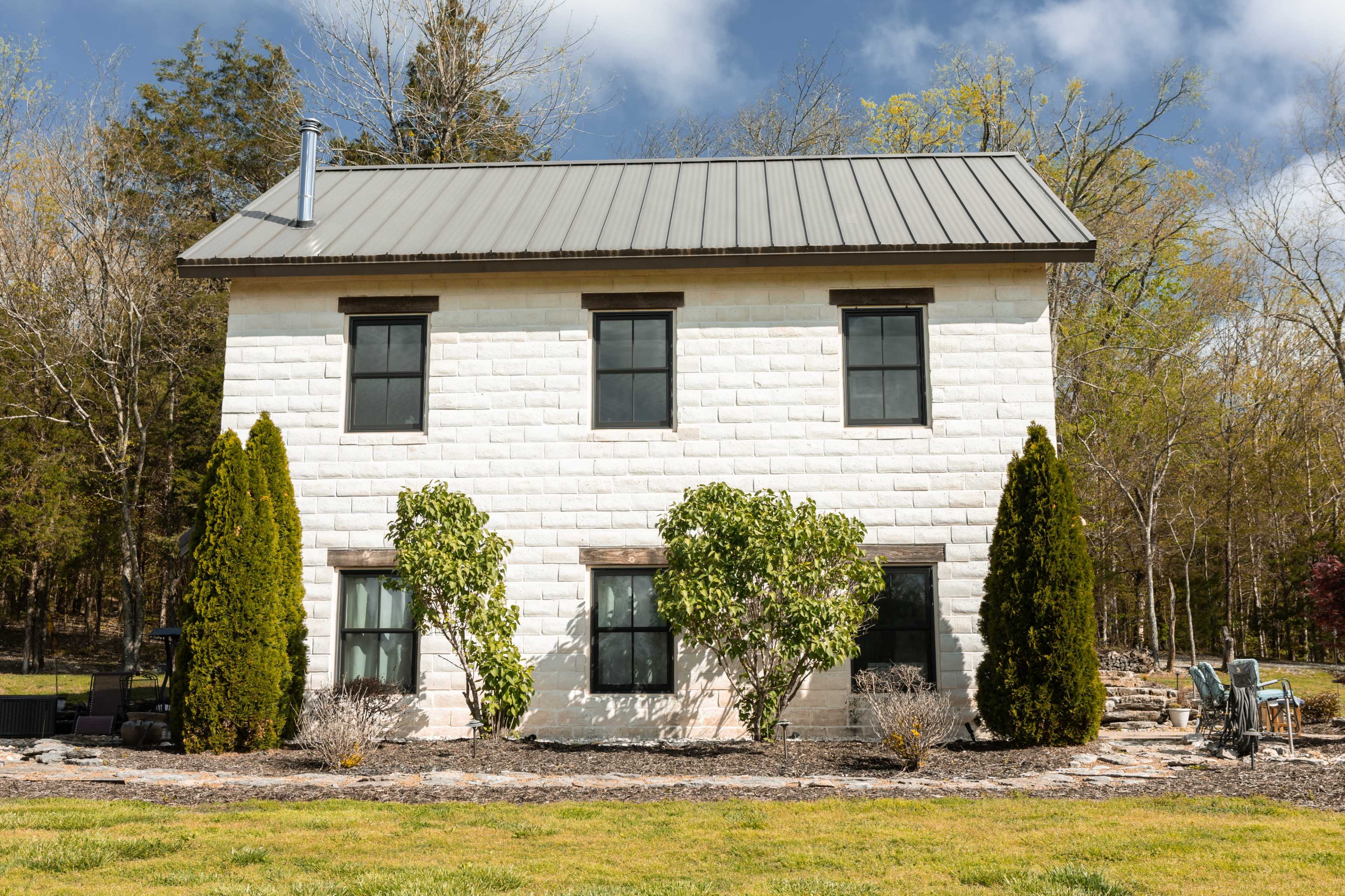 A two-story house with a metal roof and white painted block exterior is surrounded by greenery and landscaping.