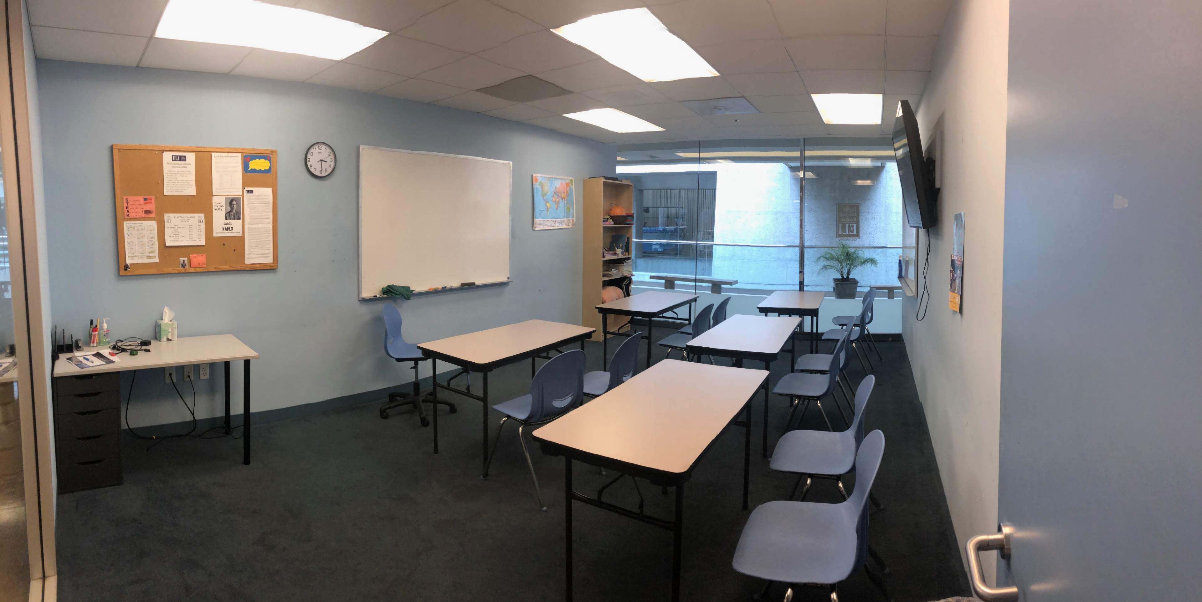 A classroom with blue walls features several tables and chairs arranged around a whiteboard and a clock on the wall.