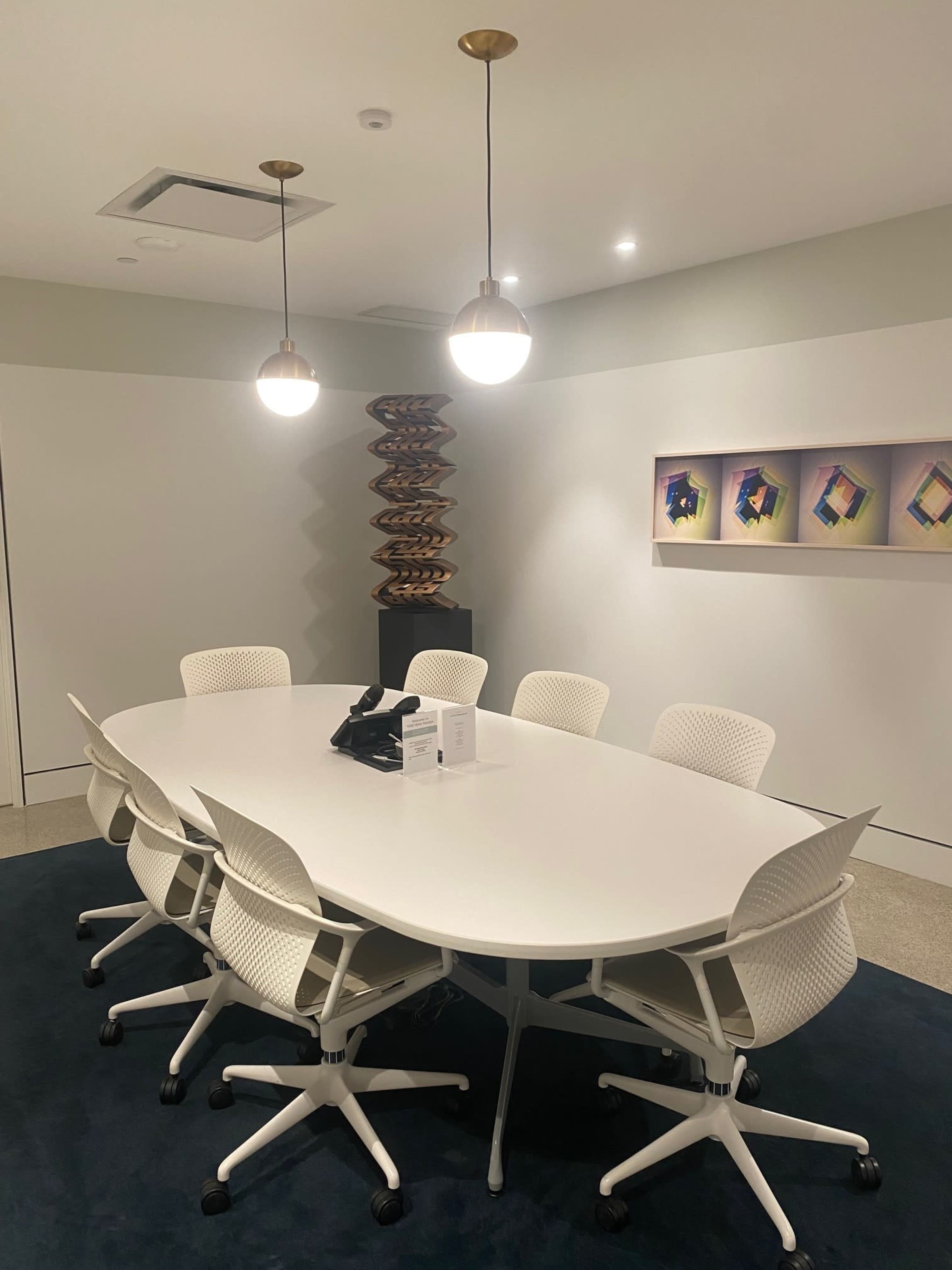 A modern conference room features an oval white table surrounded by six white chairs, with pendant lights above and a decorative sculpture in the corner.