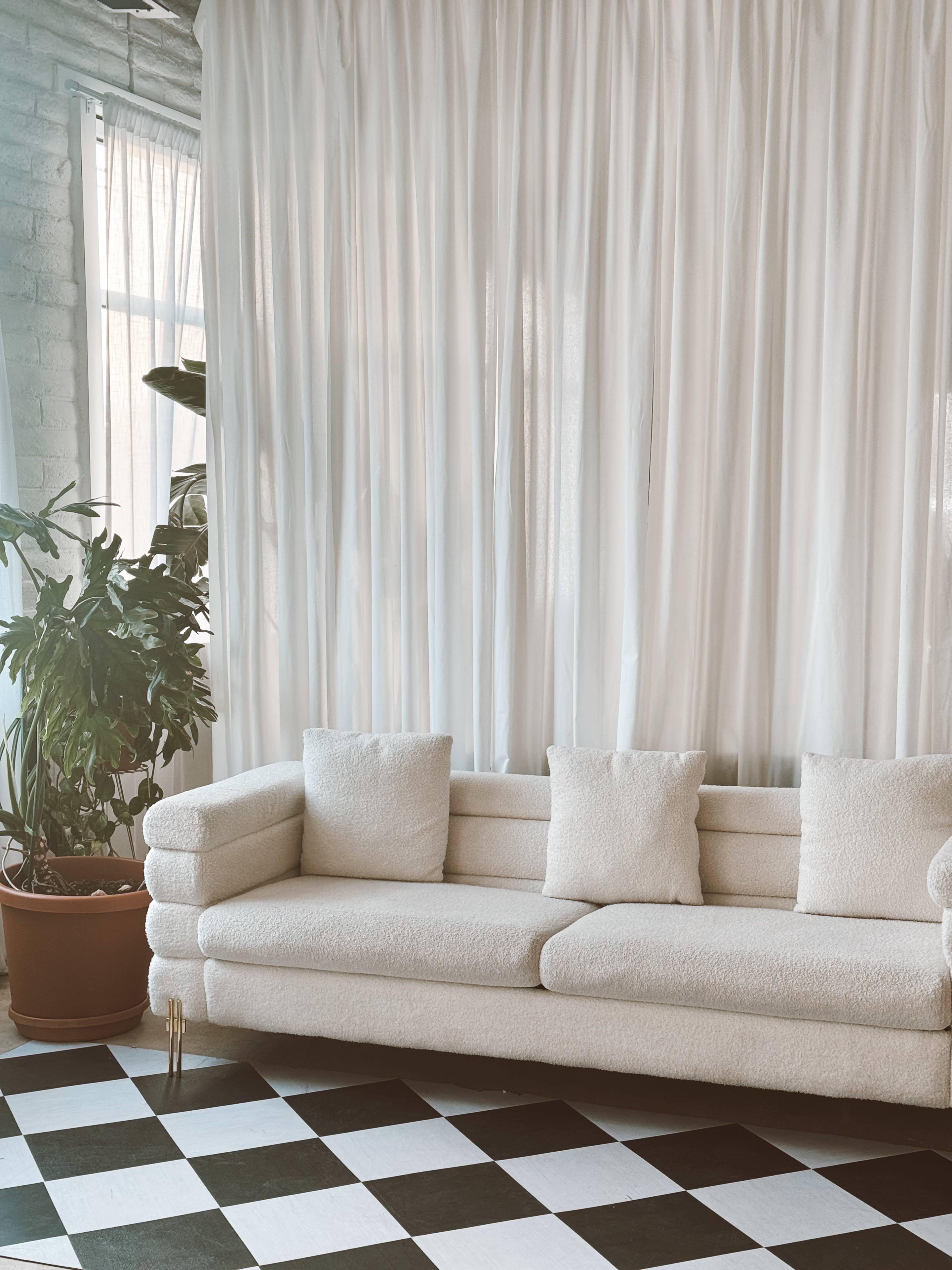 A light-colored sofa with two cushions is positioned in front of a sheer white curtain and a potted plant, on a black and white checkered floor.
