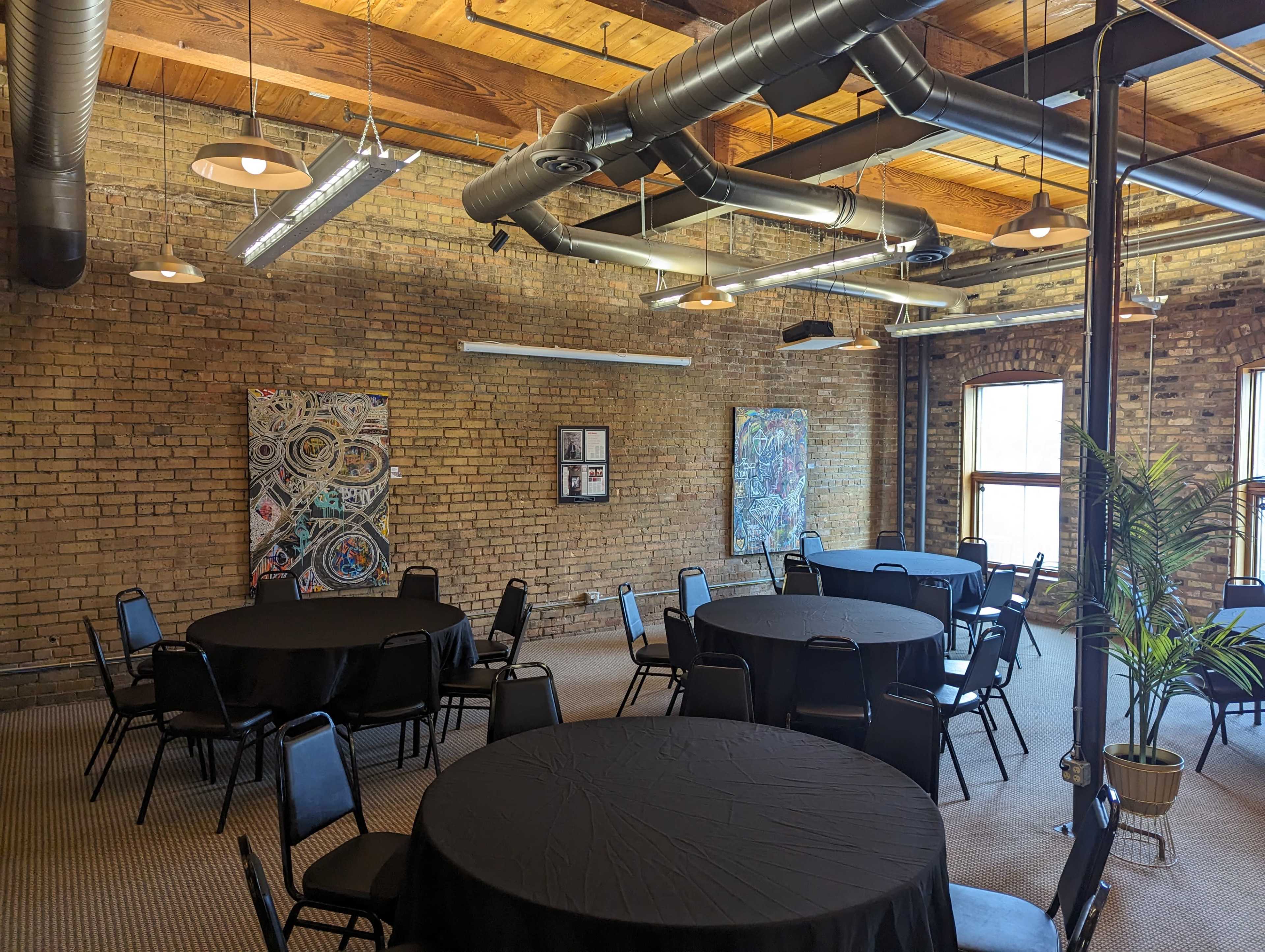 A spacious room with exposed brick walls, featuring several round tables covered with black tablecloths and artwork displayed on the walls.
