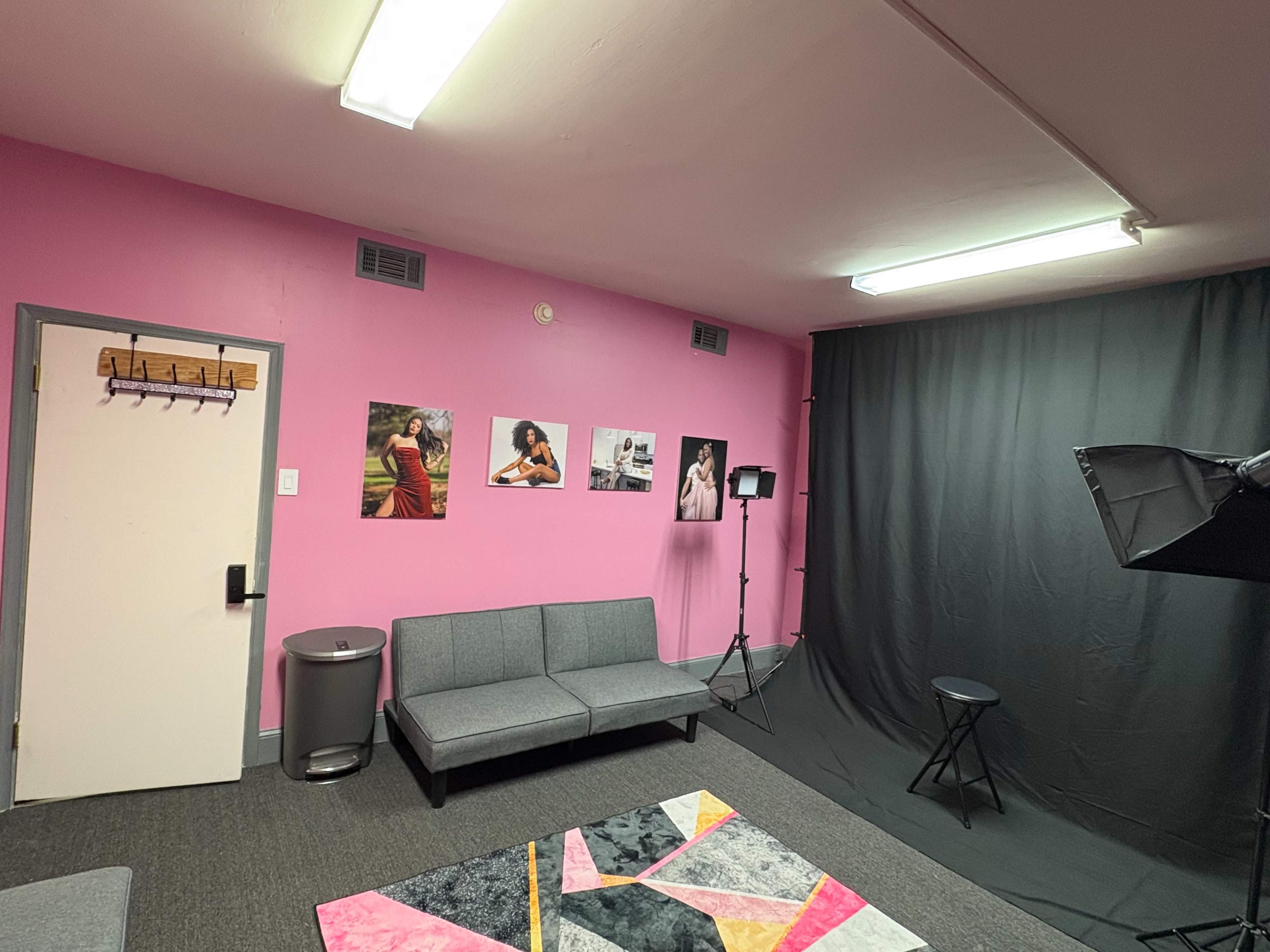 The image shows a studio room with pink walls, a gray couch, a black backdrop, and framed photographs on the wall, along with studio lighting equipment.