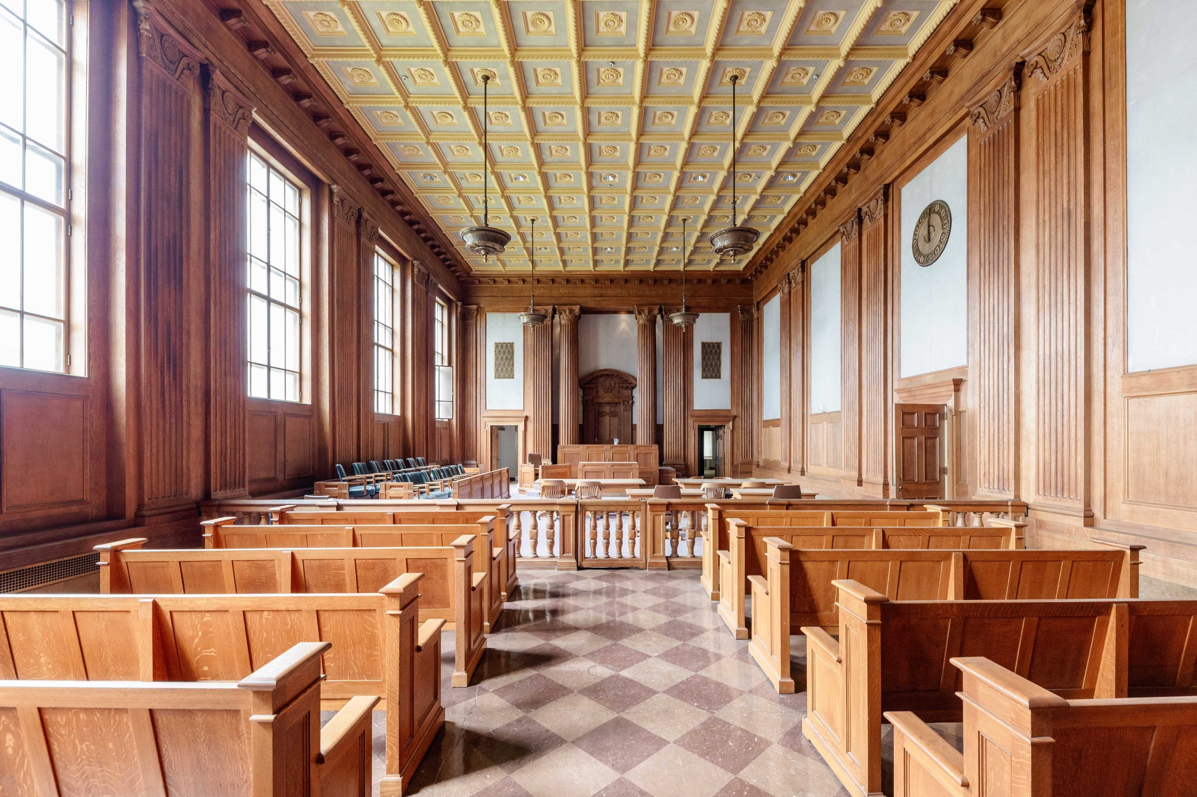 The image shows a spacious, empty courtroom with wooden benches, a judge's bench at the front, and large windows allowing natural light to enter.