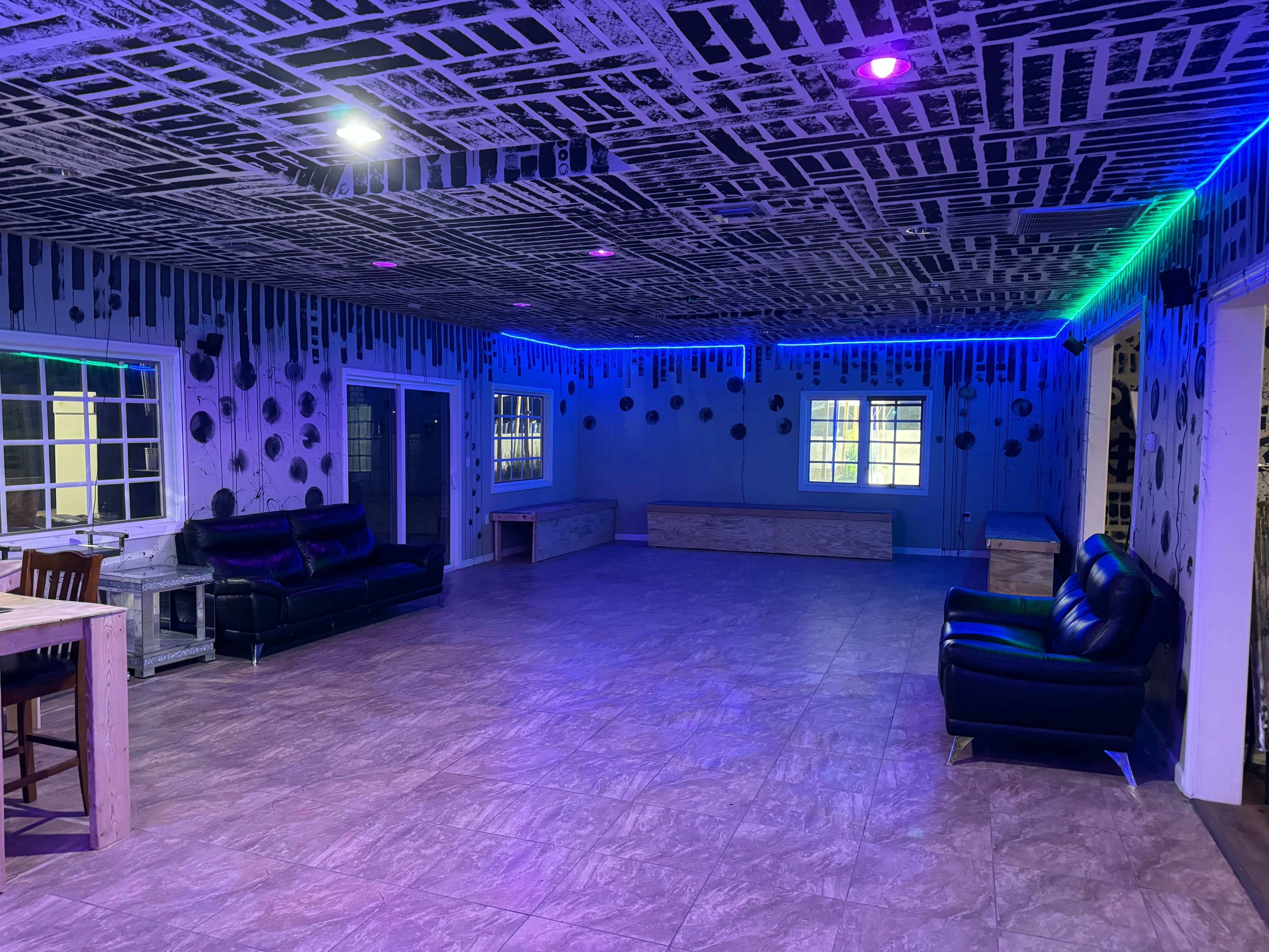 A spacious room with a patterned black and white ceiling, purple and green ambient lighting, and a few pieces of furniture, including sofas and tables.