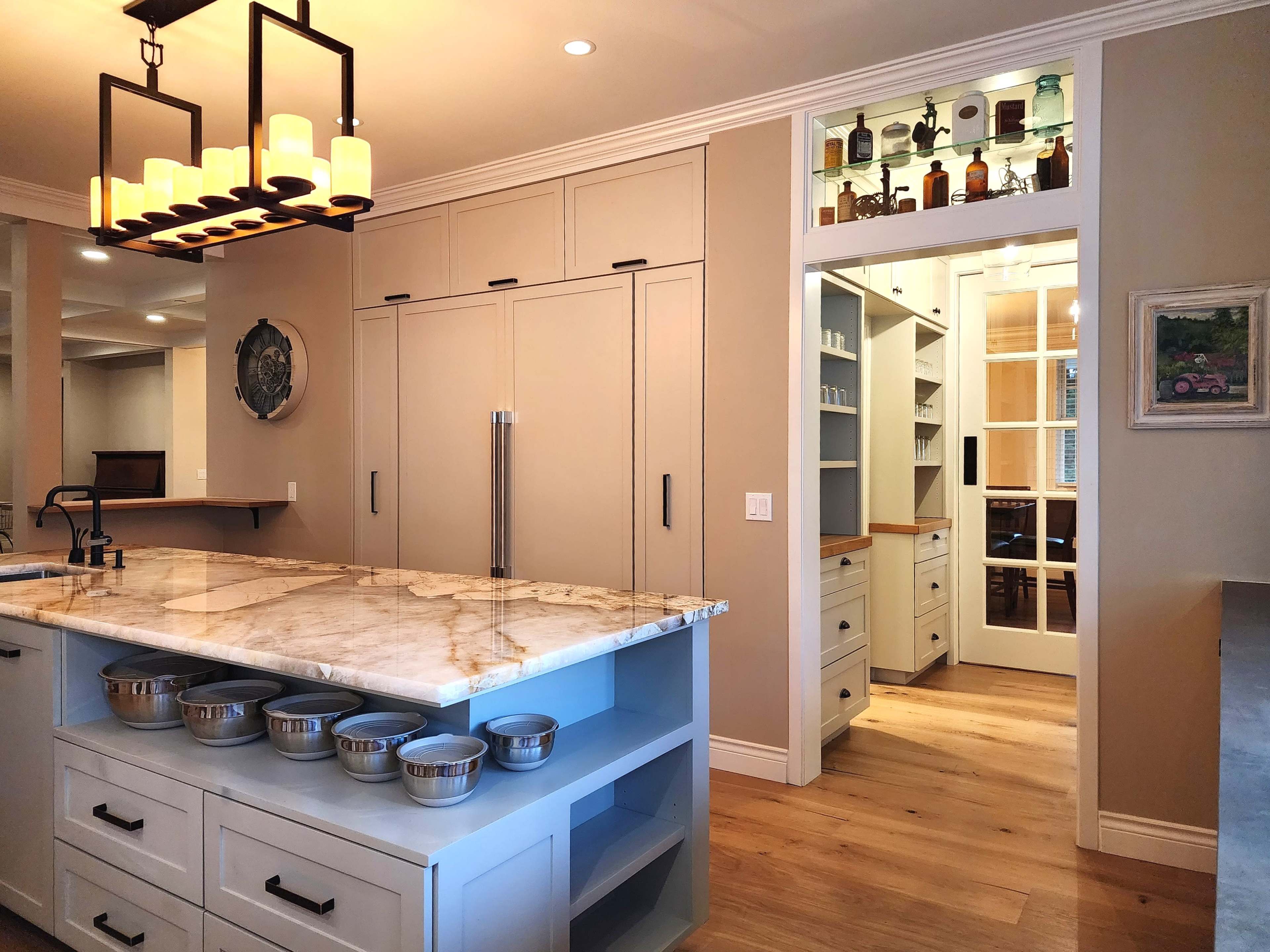 A modern kitchen features a large marble island, cabinetry with a neutral tone, and a doorway leading to a pantry space with built-in shelving.