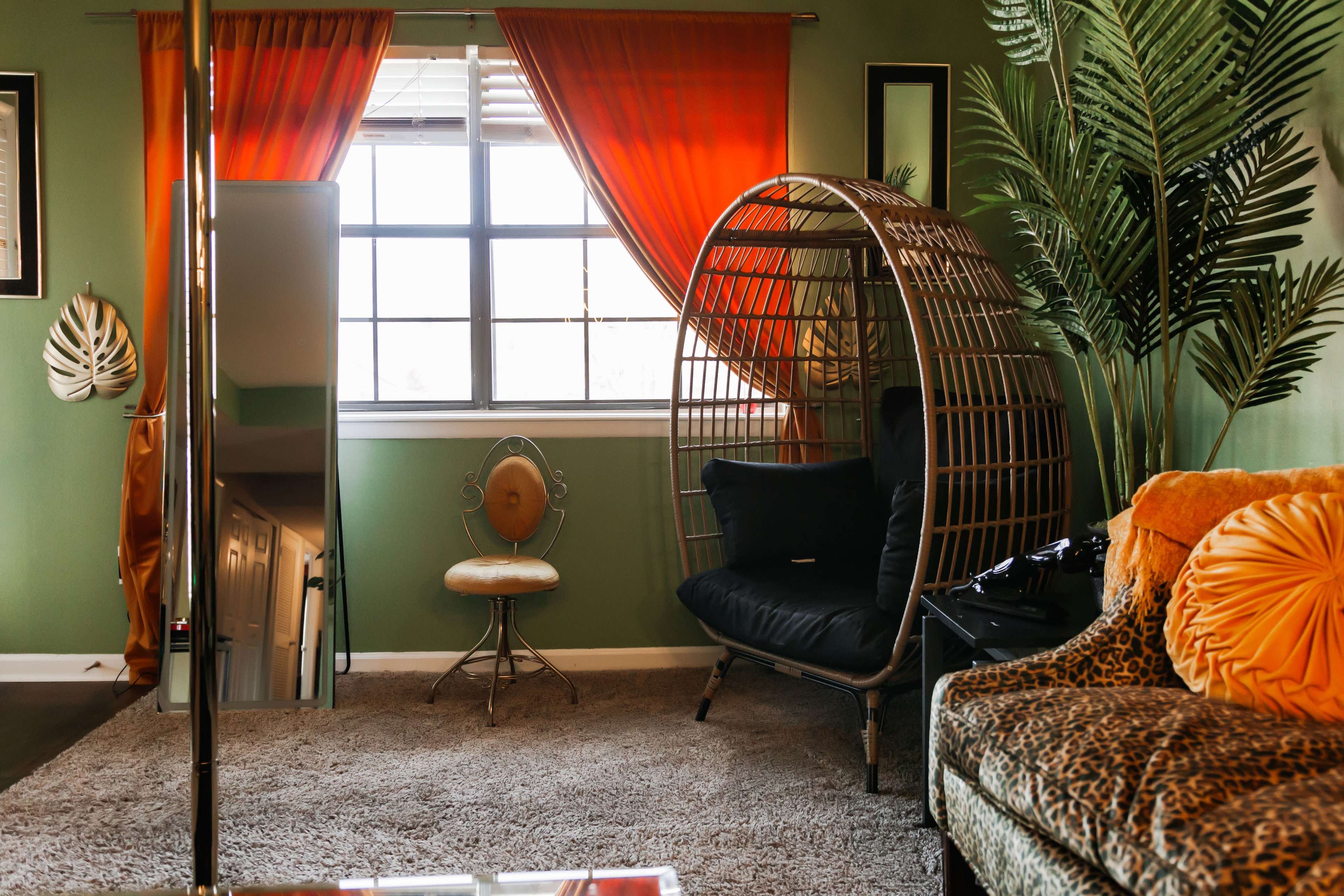 The image shows a living room with a large, round wicker chair, a small decorative chair, a mirror, and vibrant orange curtains against a green wall.