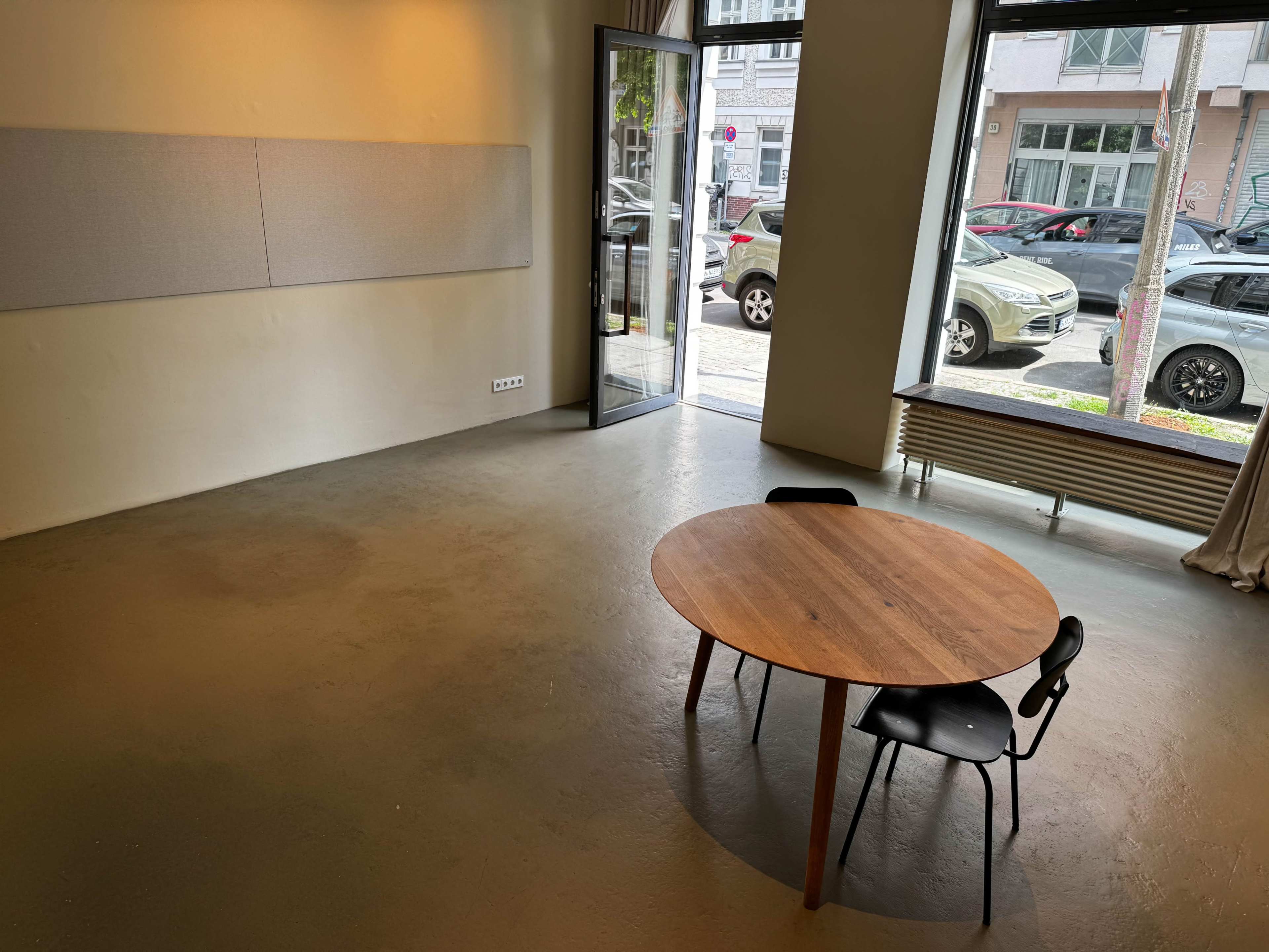 The image shows a minimalistic room with a wooden table and a black chair, featuring a large window that overlooks a street with parked cars.