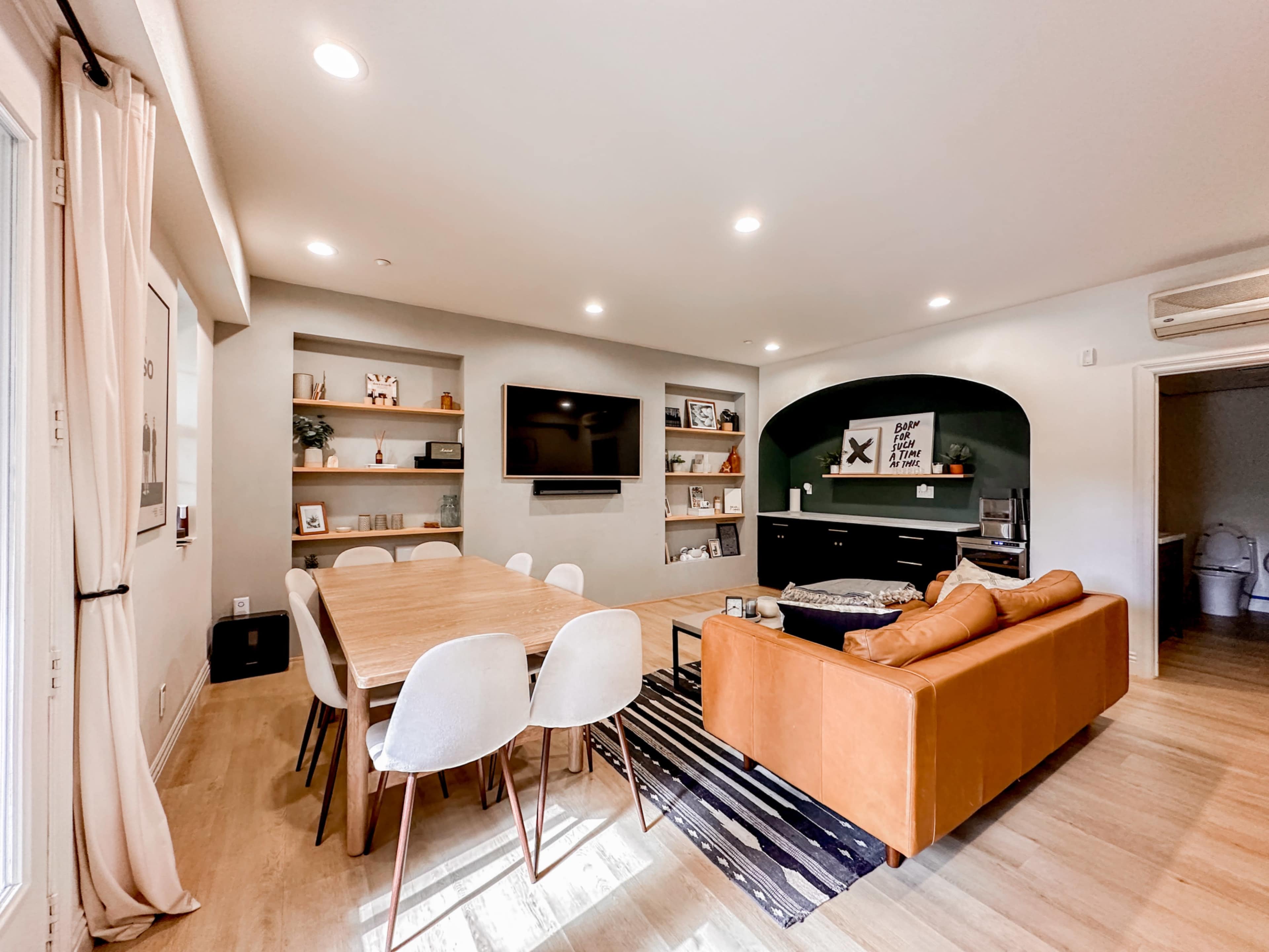 A modern living space with a dining table, a cozy sofa, shelves displaying decor, and a media center with a television.