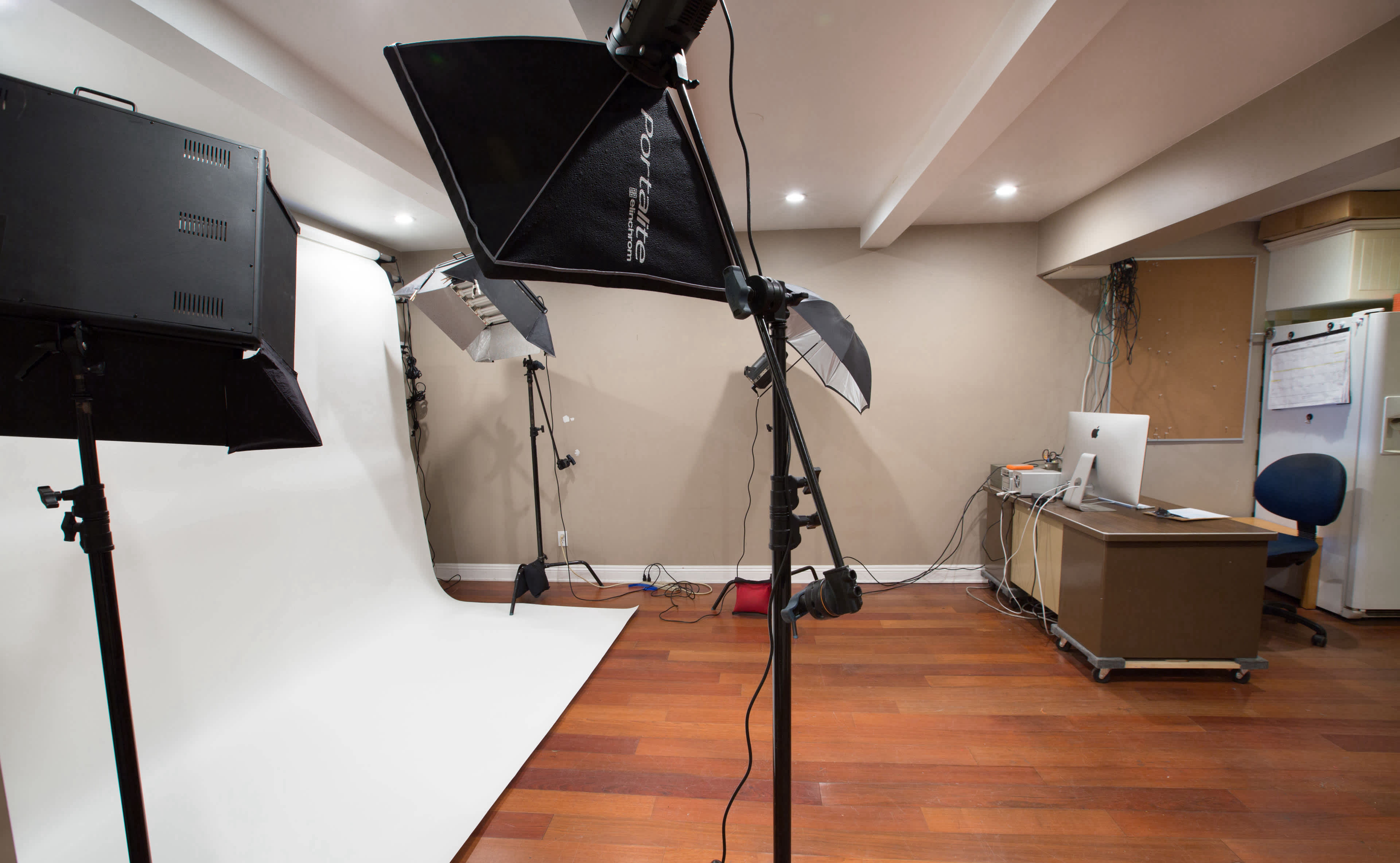 A photography studio is set up with a white backdrop, lighting equipment, and a computer workstation in a room with wooden flooring.
