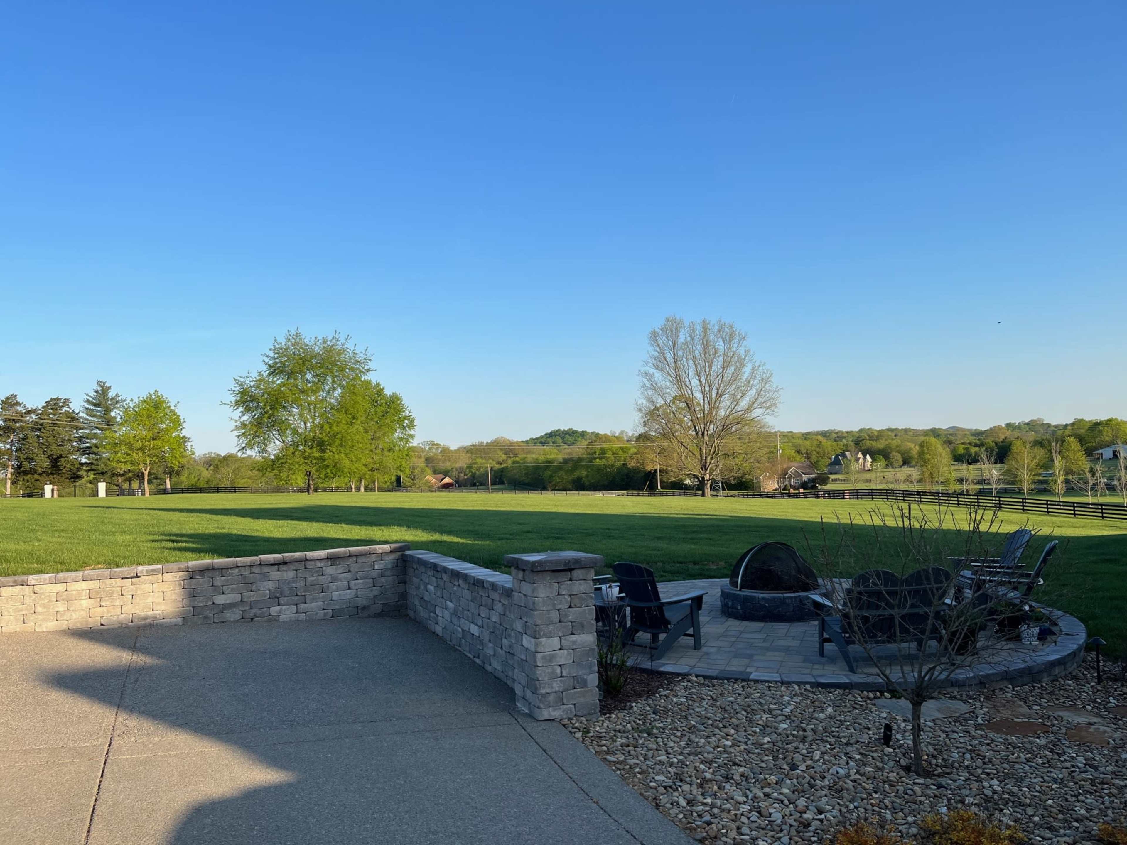 A stone patio with a fire pit and wooden chairs overlooks a grassy landscape and distant hills under a clear blue sky.