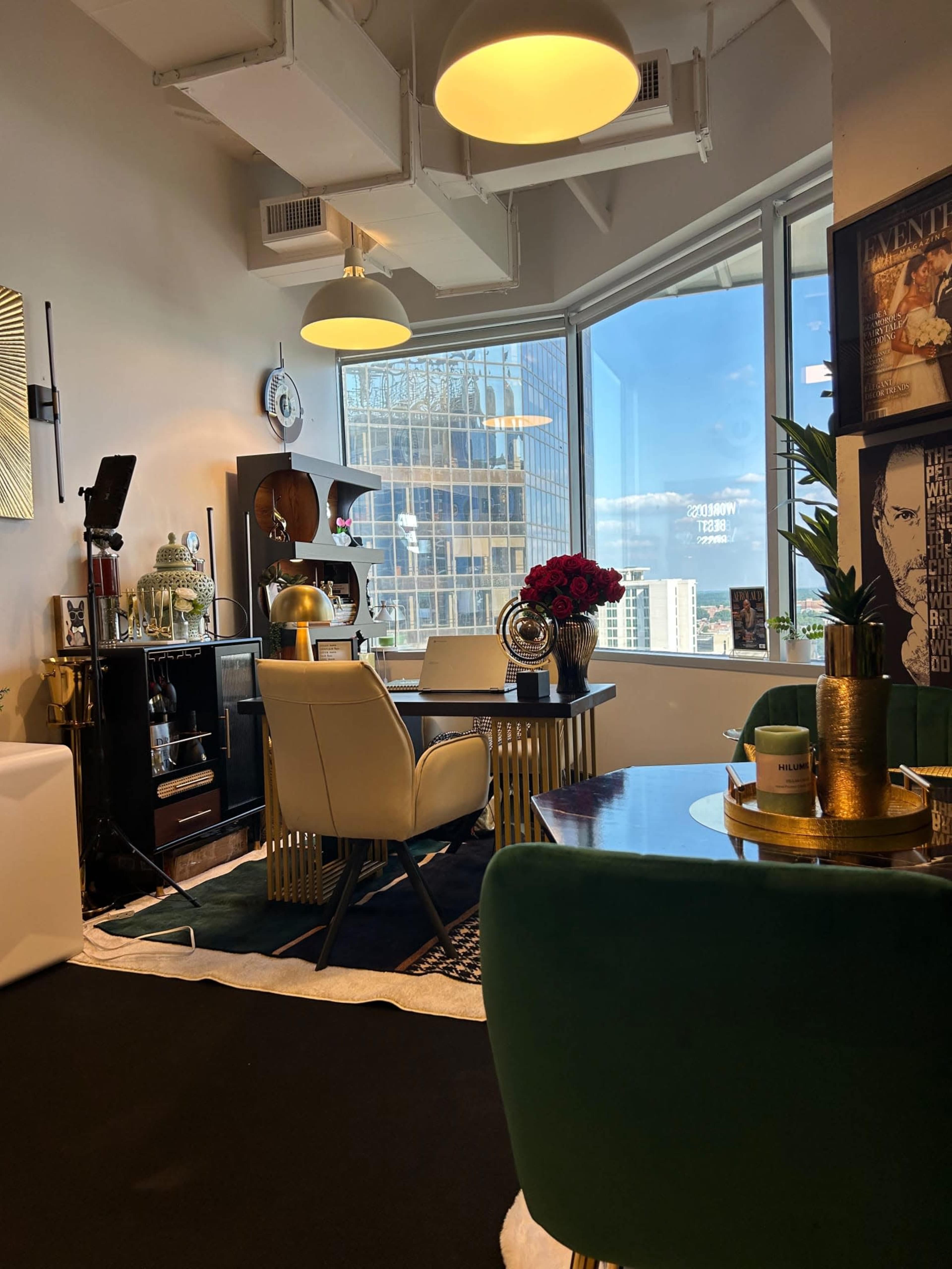 The image shows a stylish office space with modern furniture, including a round table and chairs, large windows offering a city view, and decorative plants.