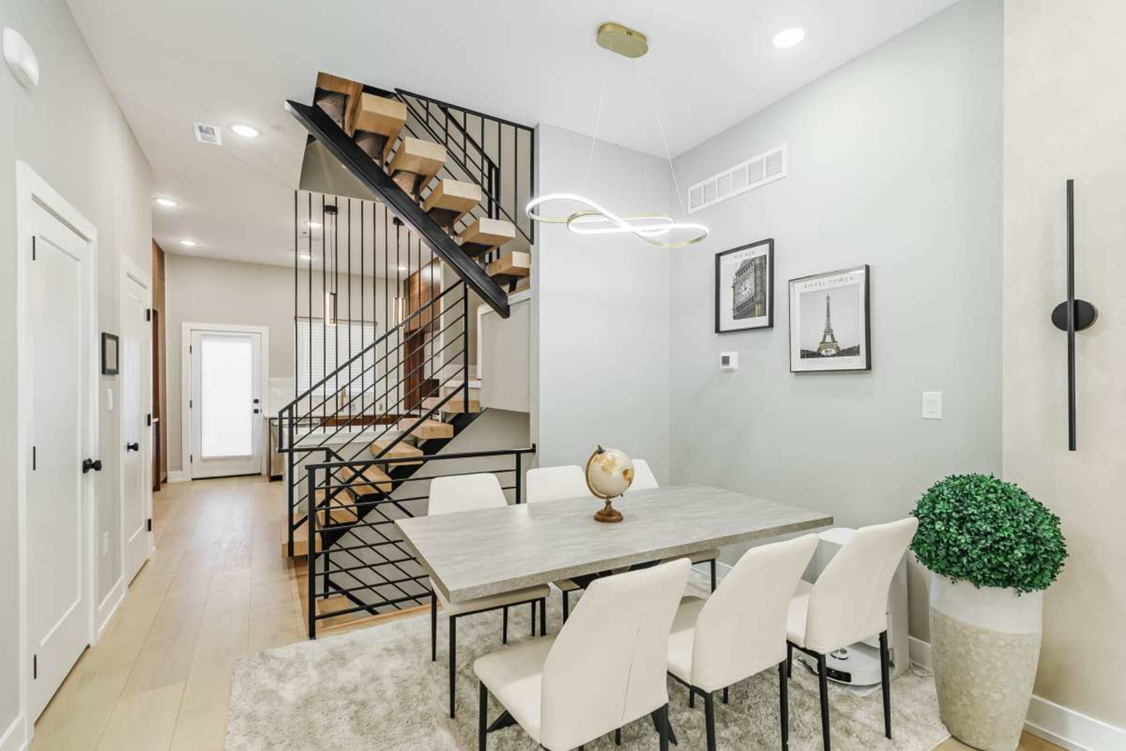A modern dining area features a large table surrounded by white chairs, with a staircase in the background and decor on the walls.