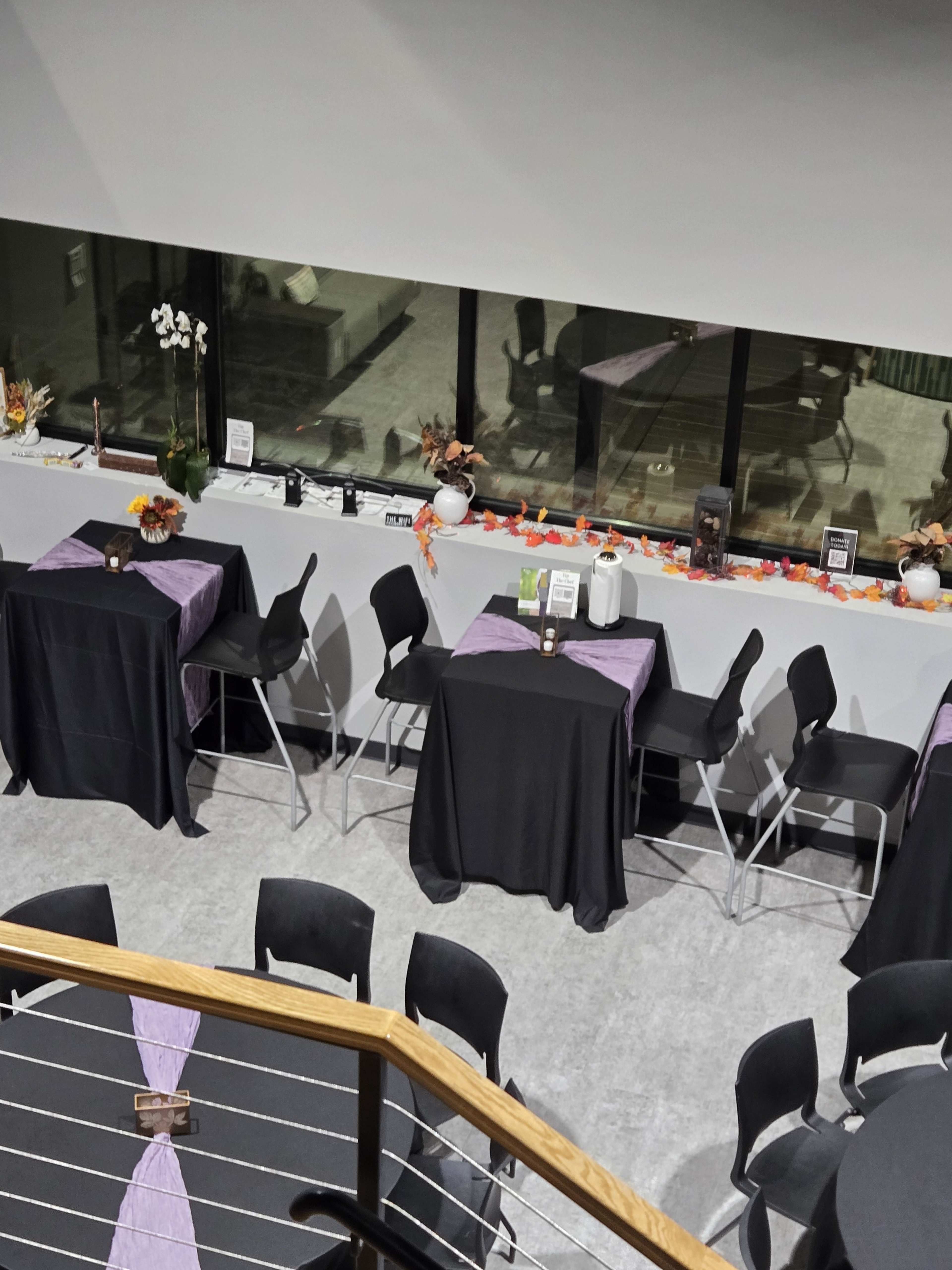 The image shows a banquet space with several round and rectangular tables covered in black cloth, each adorned with a purple accent, and decorated with fall-themed arrangements along the windowsill.