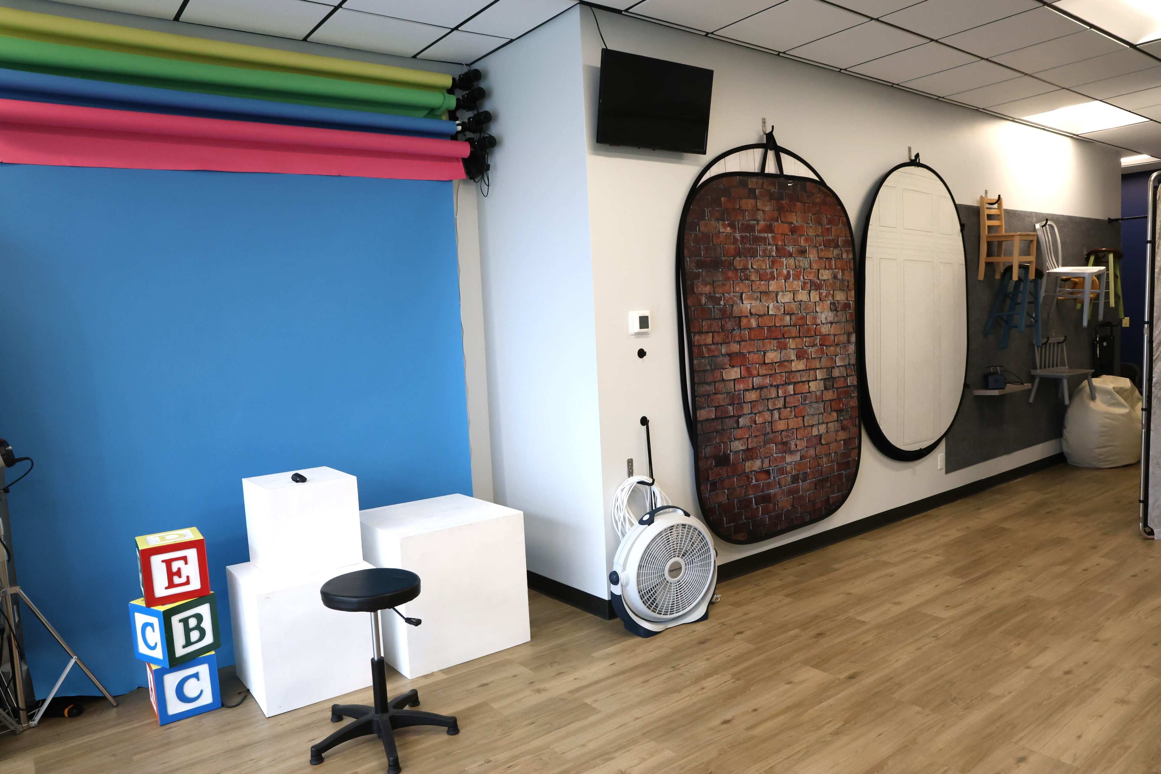 A studio space with a blue backdrop, colorful paper rolls, a fan, white props, and a brick-patterned wall display.