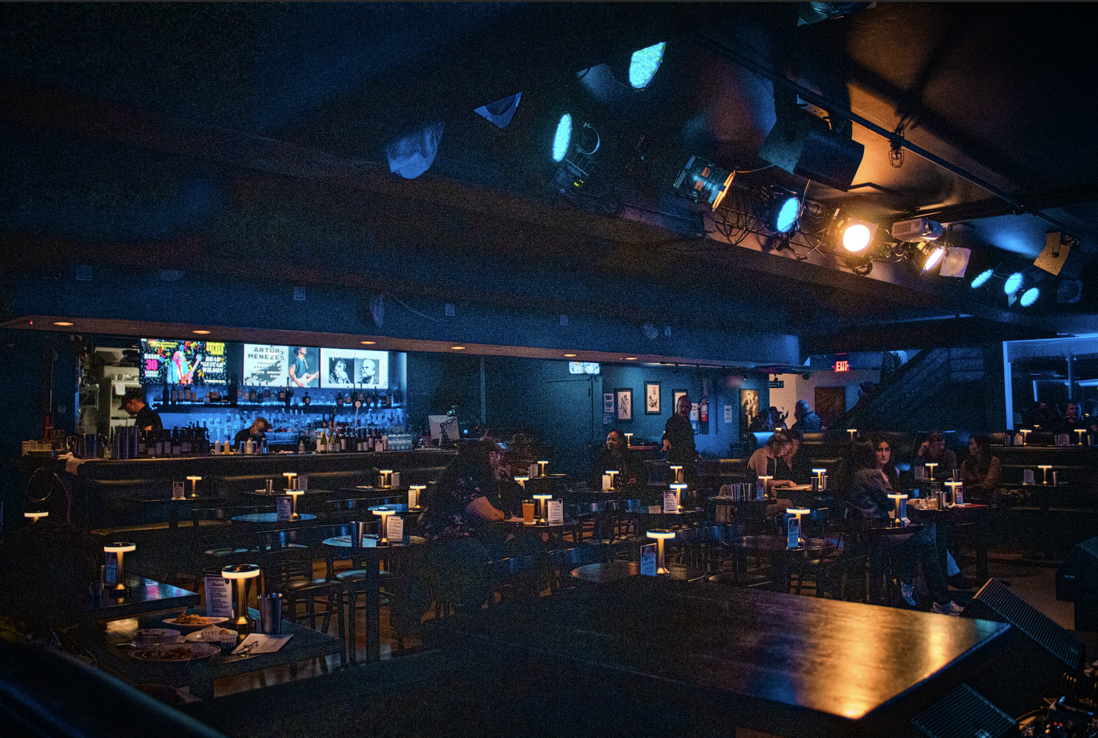 A dimly lit comedy club features rows of tables with small candles on them and a bar in the background displaying colorful screens.