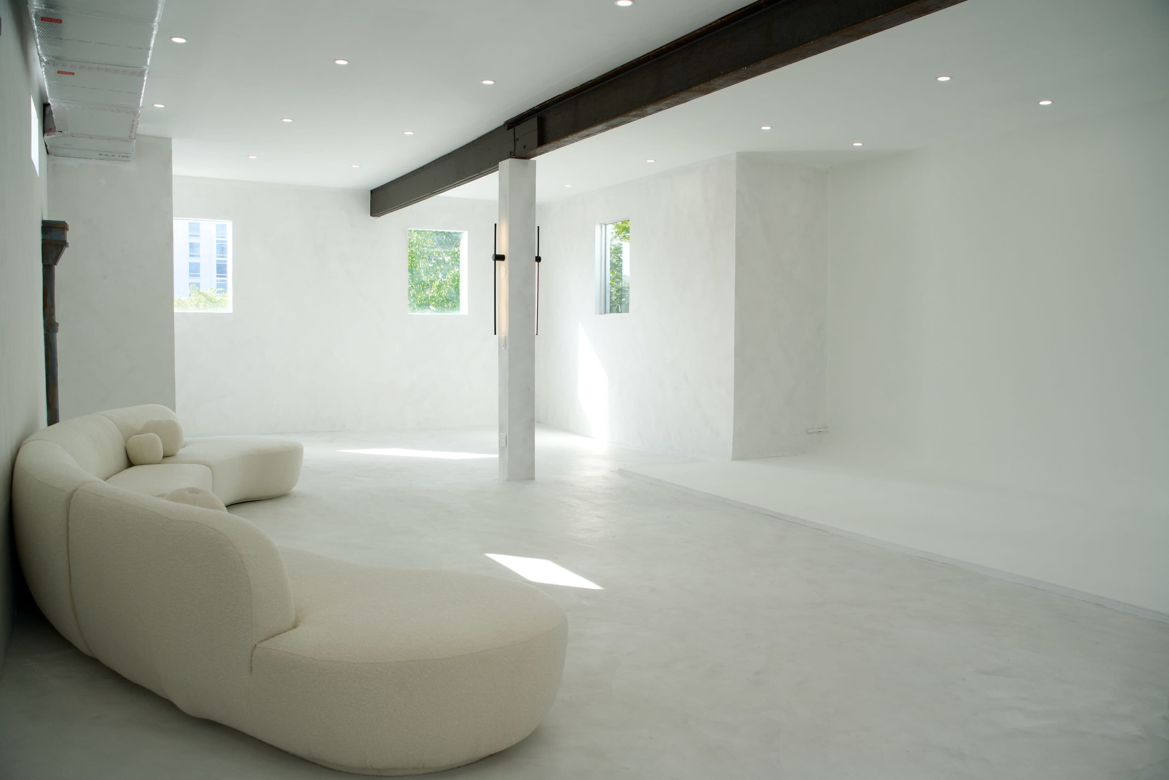 The image shows a spacious, minimally furnished room with white walls, a curved sofa, and large windows allowing natural light to enter.