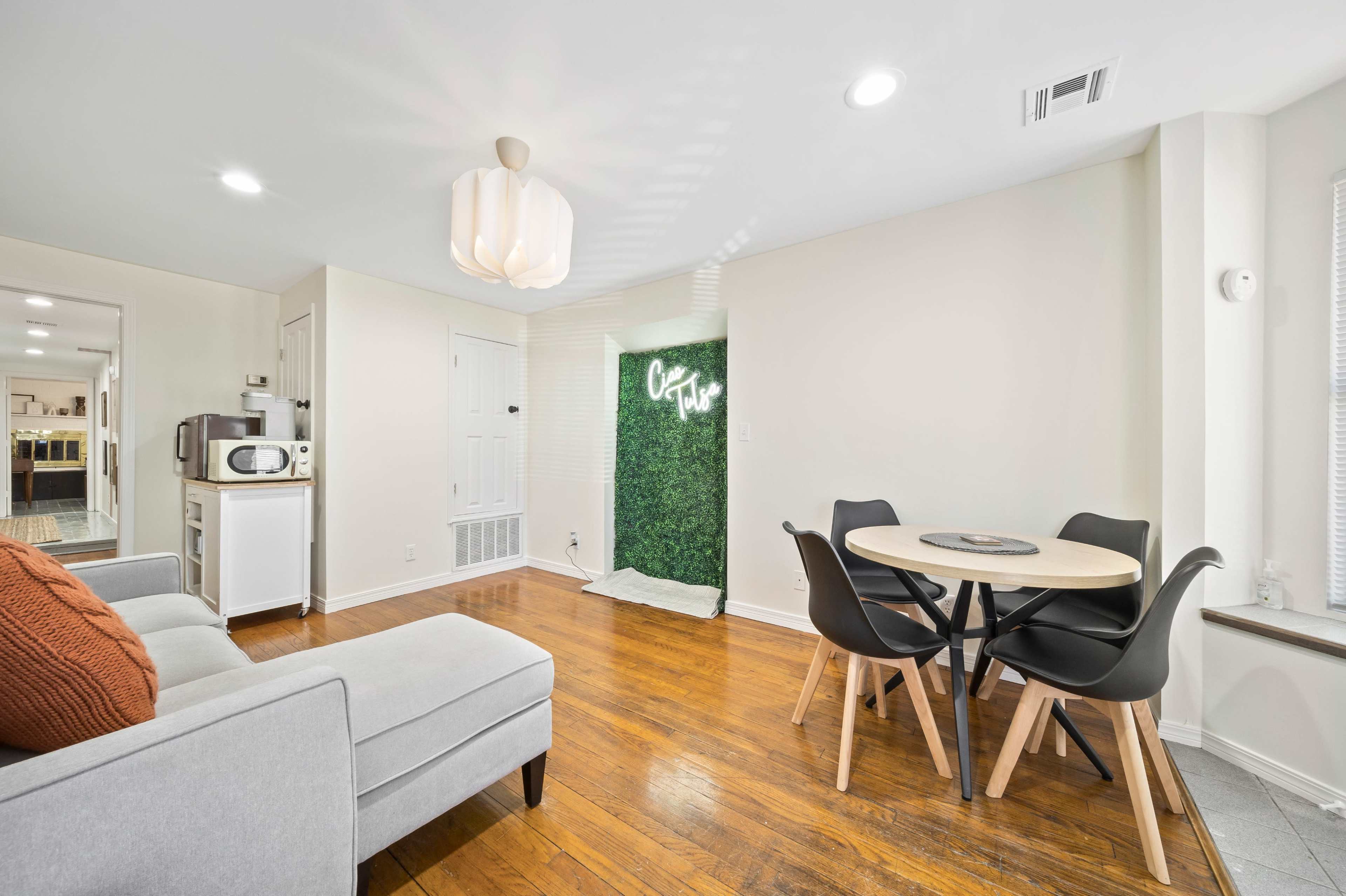 The image shows a cozy living area with a gray sofa, a circular dining table, and a decorative green wall feature with illuminated writing.