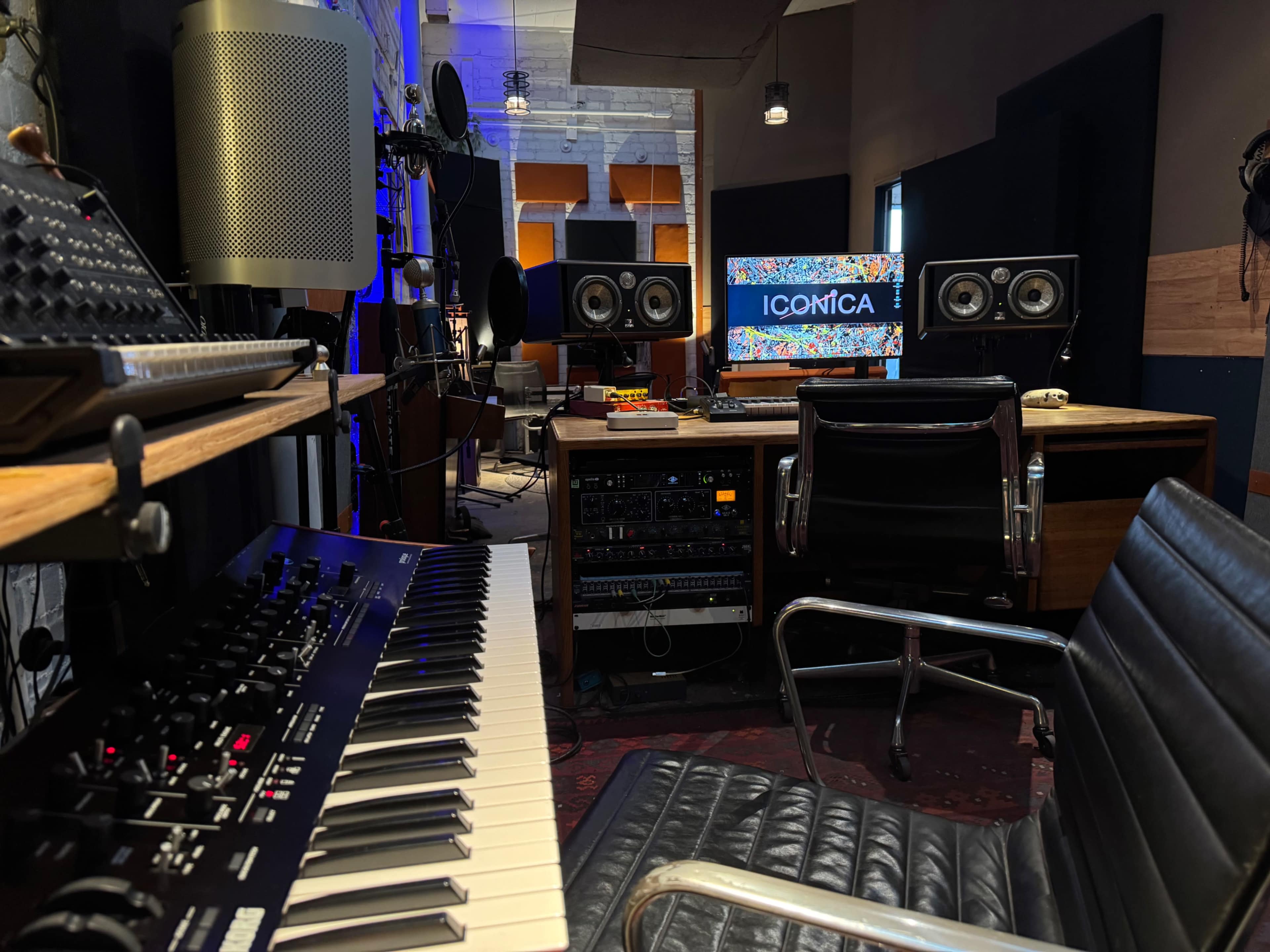 A music studio setup features a keyboard in the foreground, speakers on either side of a computer, and various audio equipment arranged on a wooden desk.