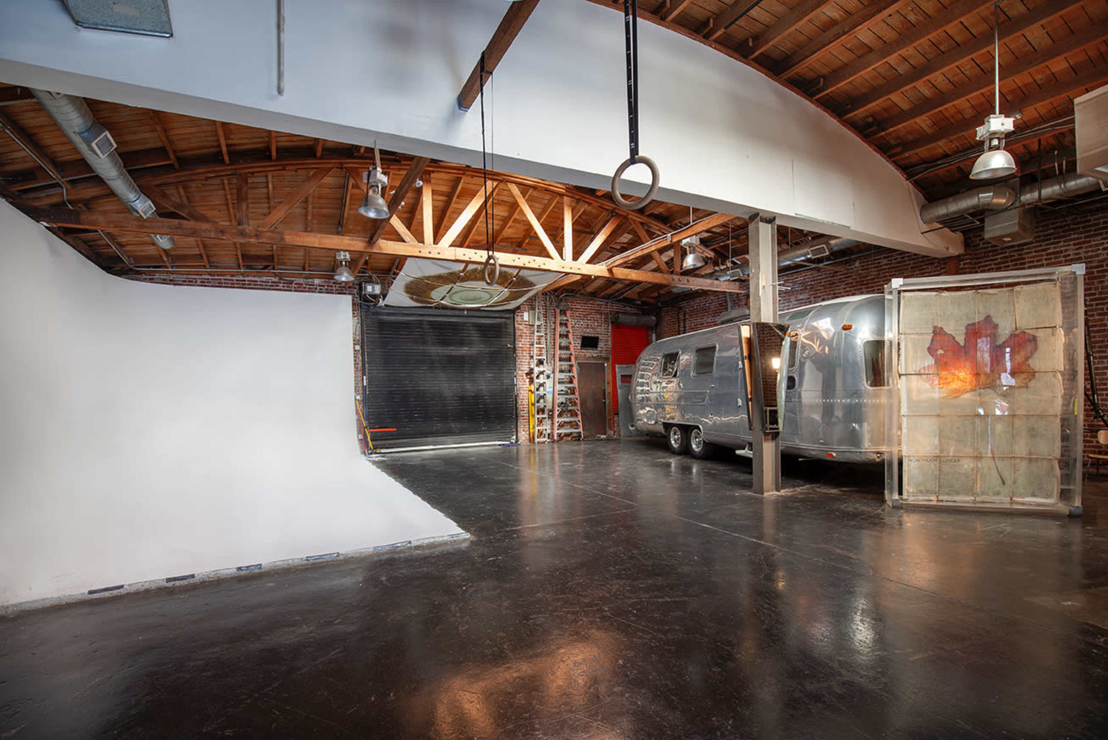 An industrial-style space with a polished black floor, exposed wooden beams, a large white backdrop, an Airstream trailer, and a garage door at the rear.