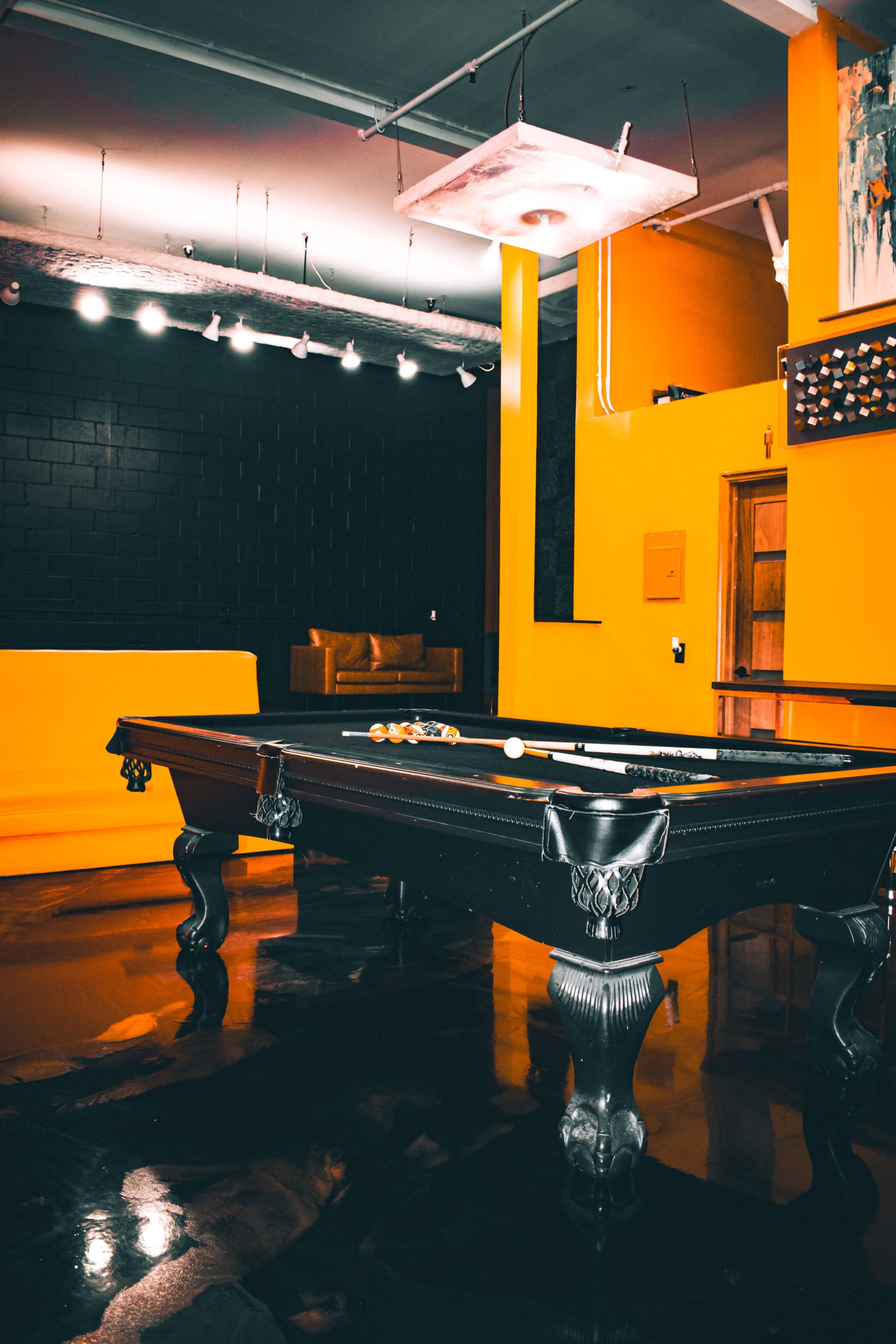 A black billiard table sits in a room with yellow and black walls, featuring industrial lighting and a polished floor.