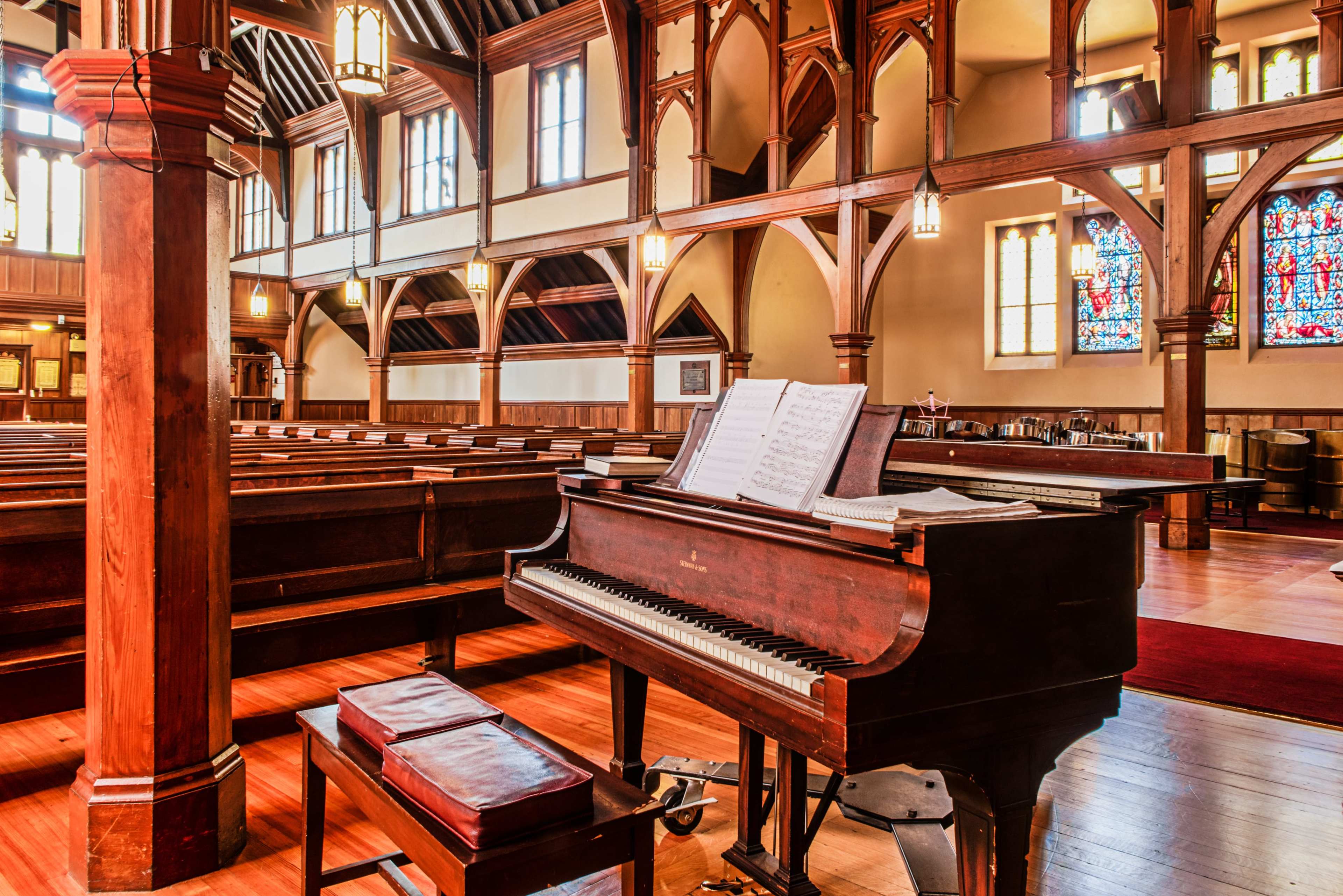 The interior of a church features wooden pews and a grand piano with sheet music, illuminated by stained glass windows.