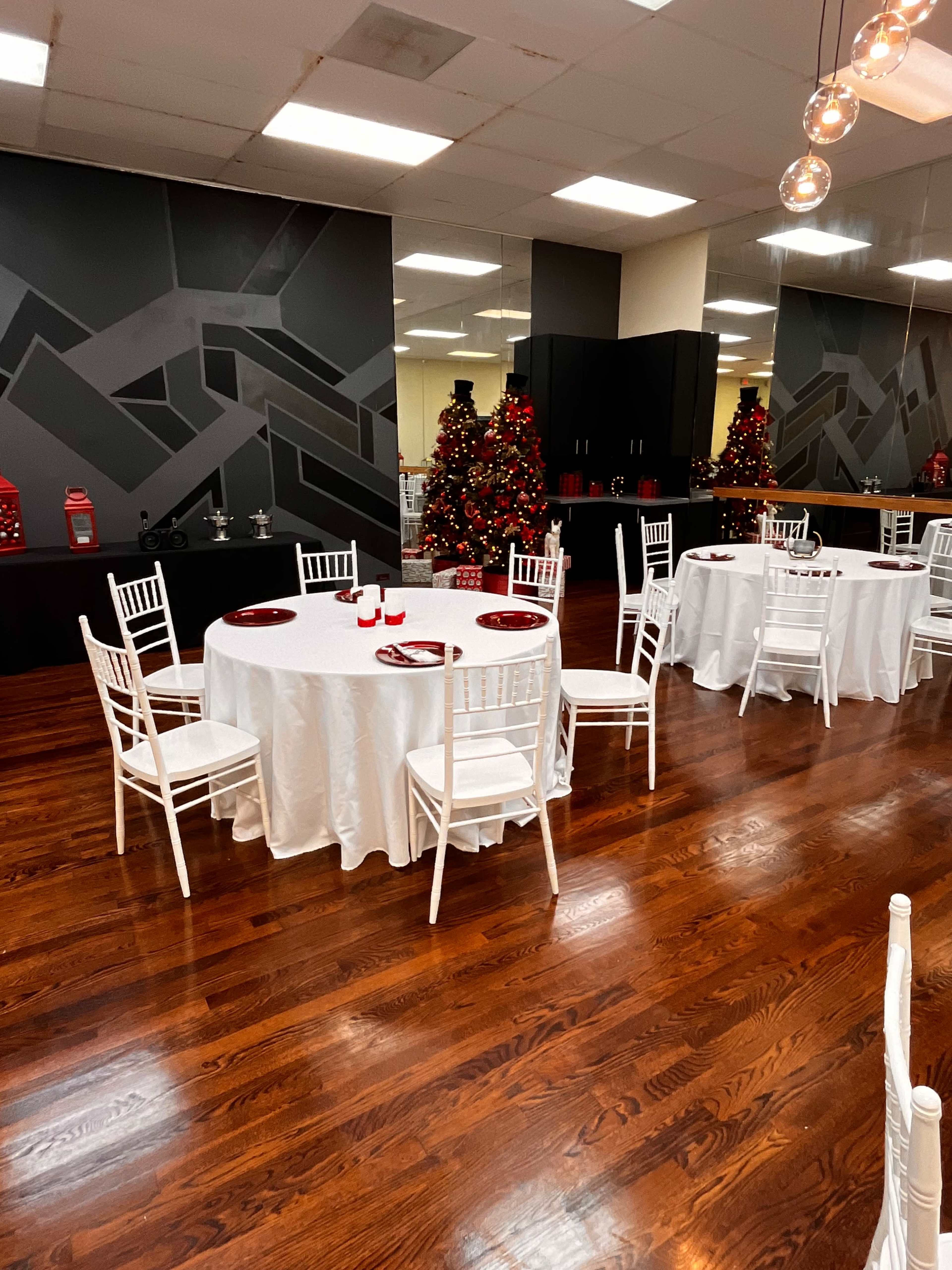 The image shows a spacious banquet room with white round tables set with red plates and surrounded by white chairs, decorated with Christmas trees.