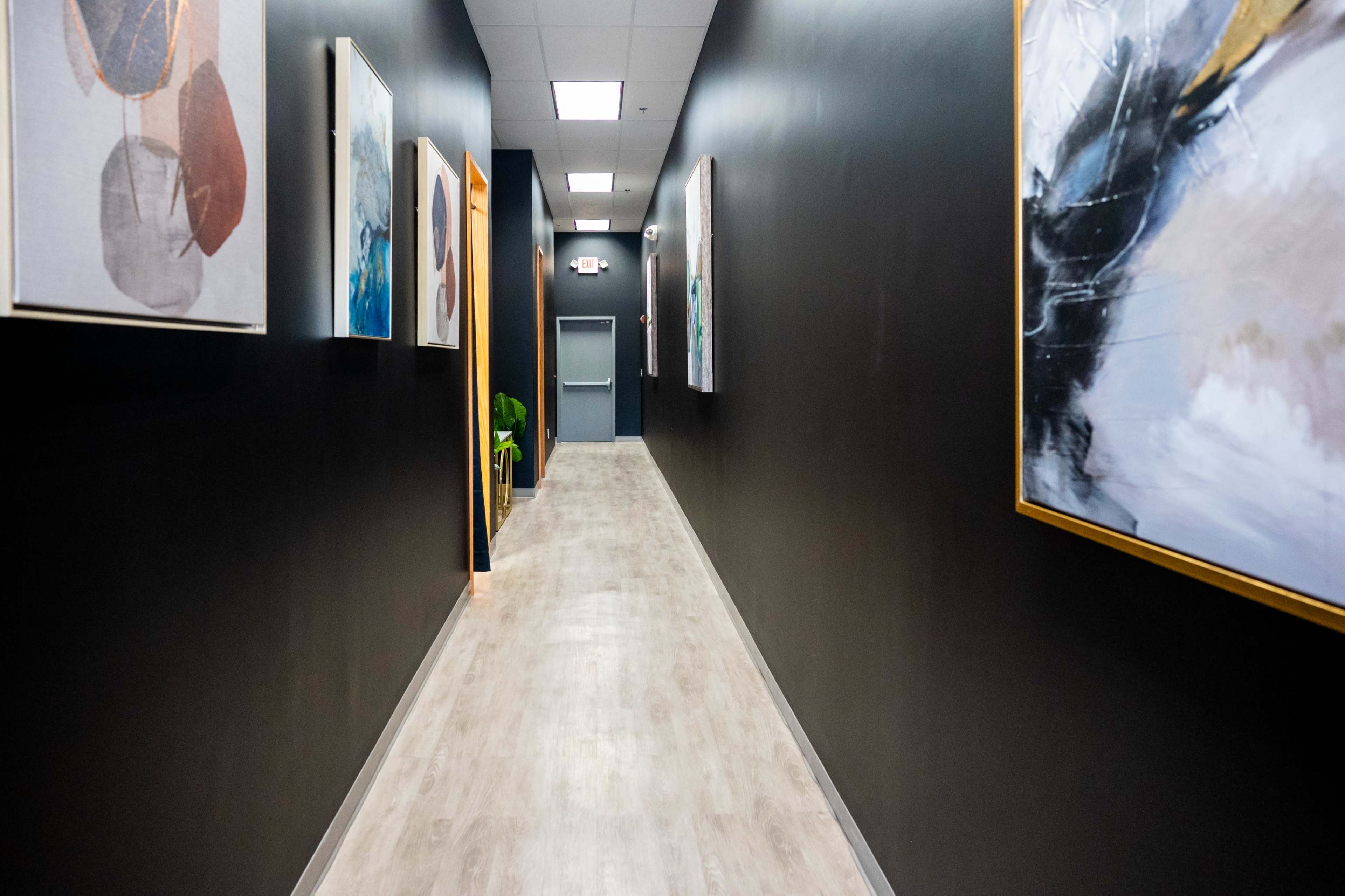A narrow hallway with dark walls, wooden flooring, and various artworks hanging on the walls.
