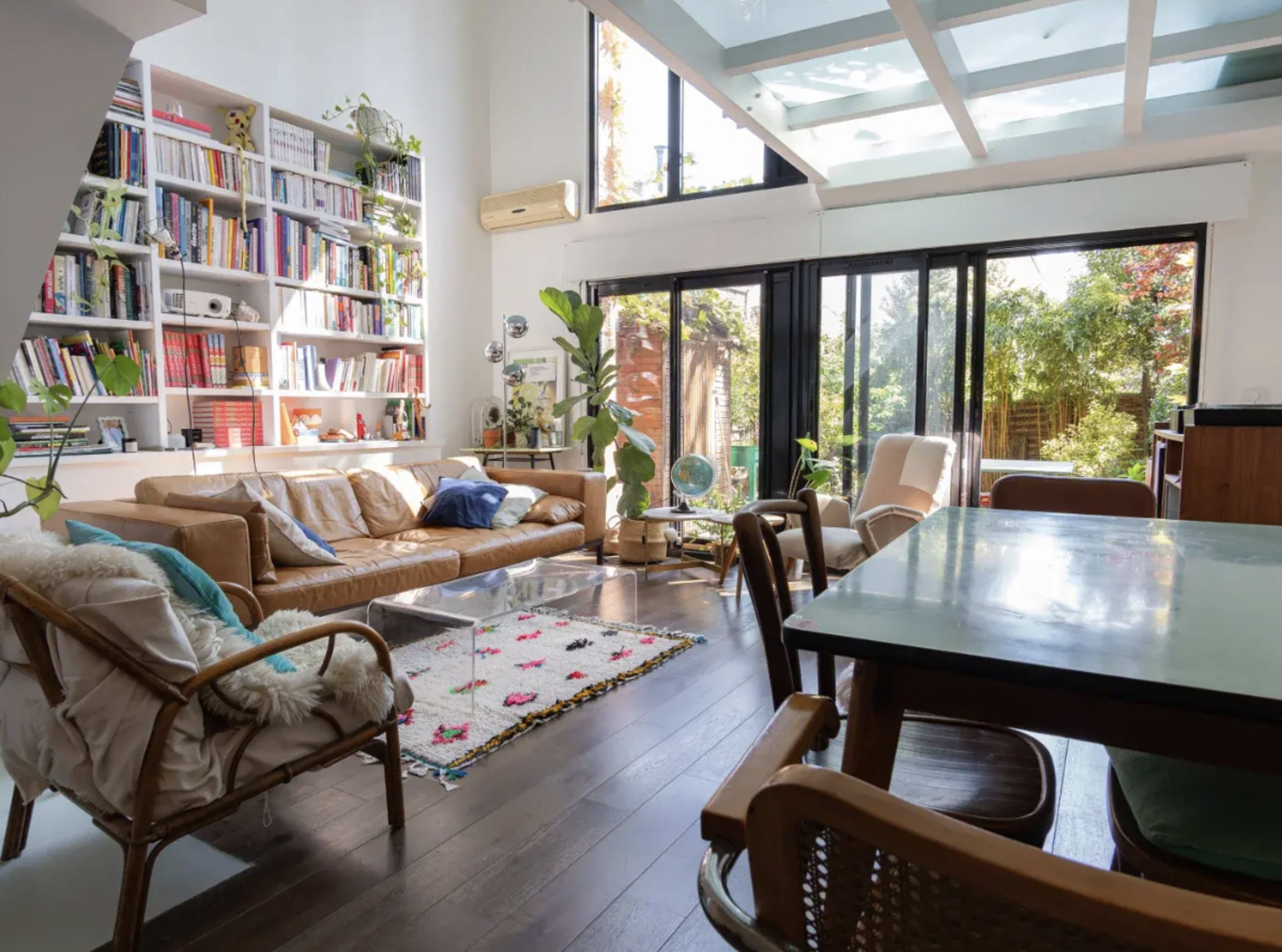 The image shows a bright, open living space with a leather sofa, a glass coffee table, and a dining area featuring a wooden table and stylish chairs.