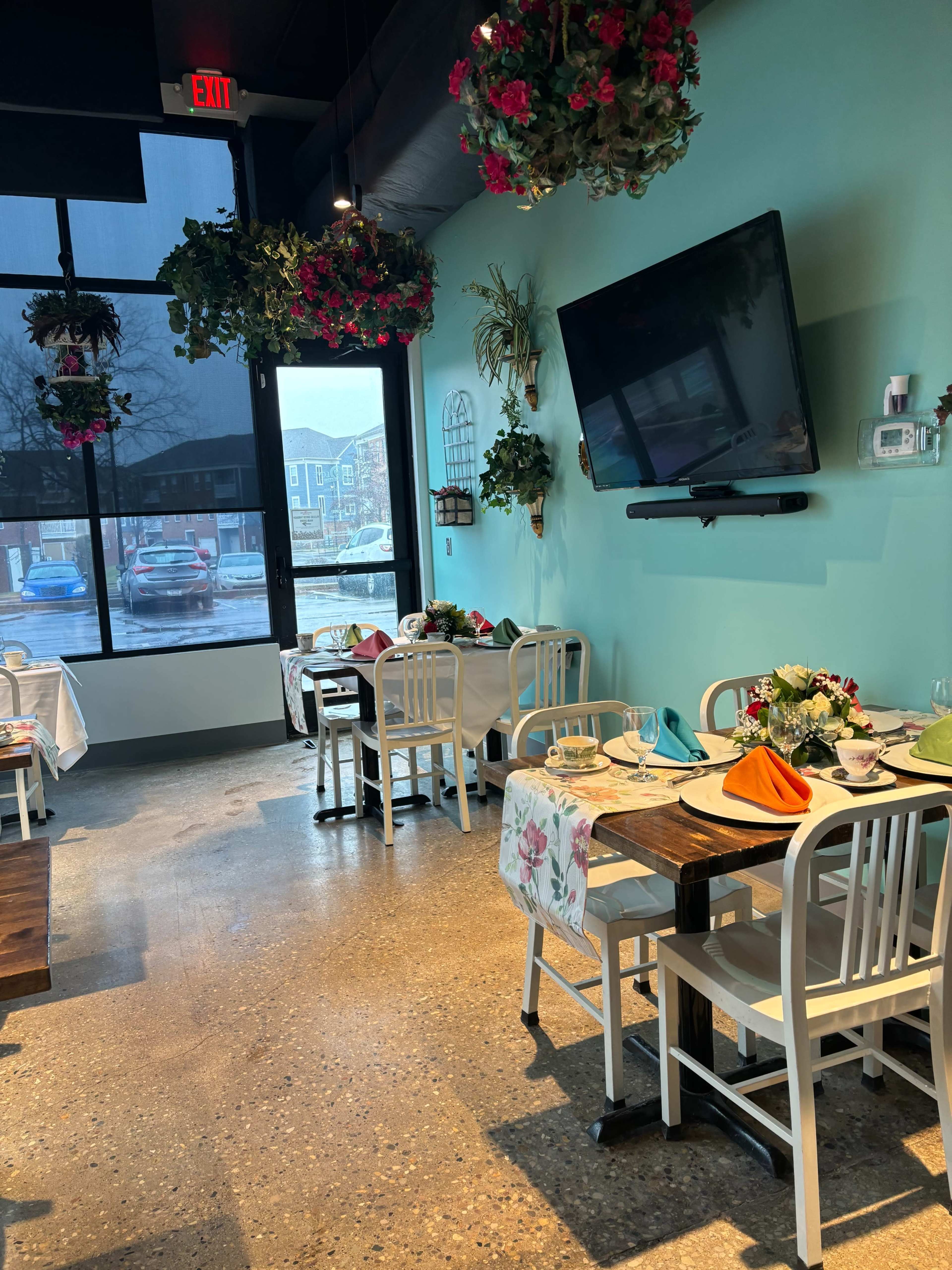 A brightly lit restaurant interior with wooden tables and white chairs, adorned with colorful table settings and hanging flower baskets.