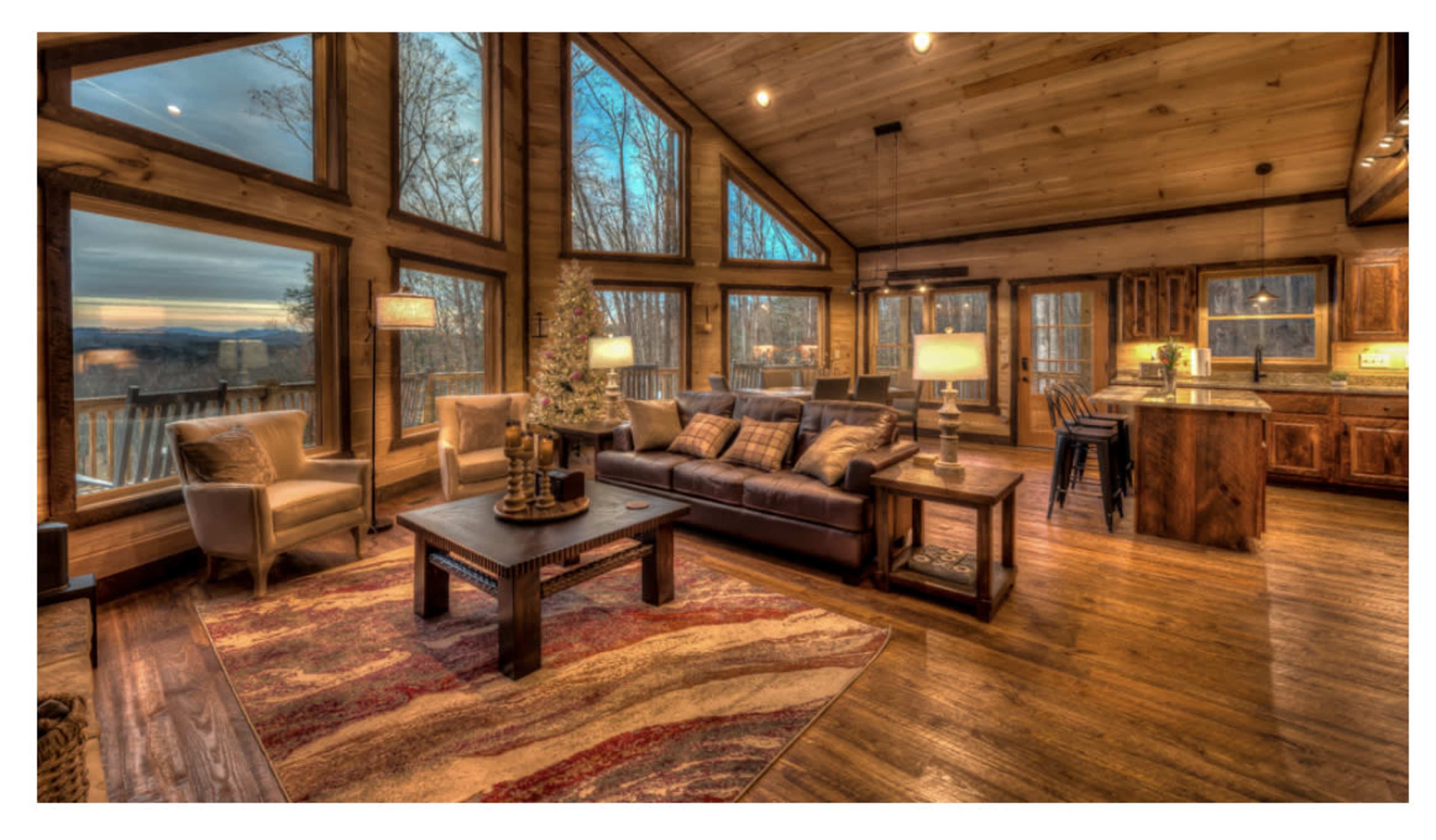 A wooden living room with large windows overlooking a wooded landscape, featuring a cozy seating area and a kitchen space.