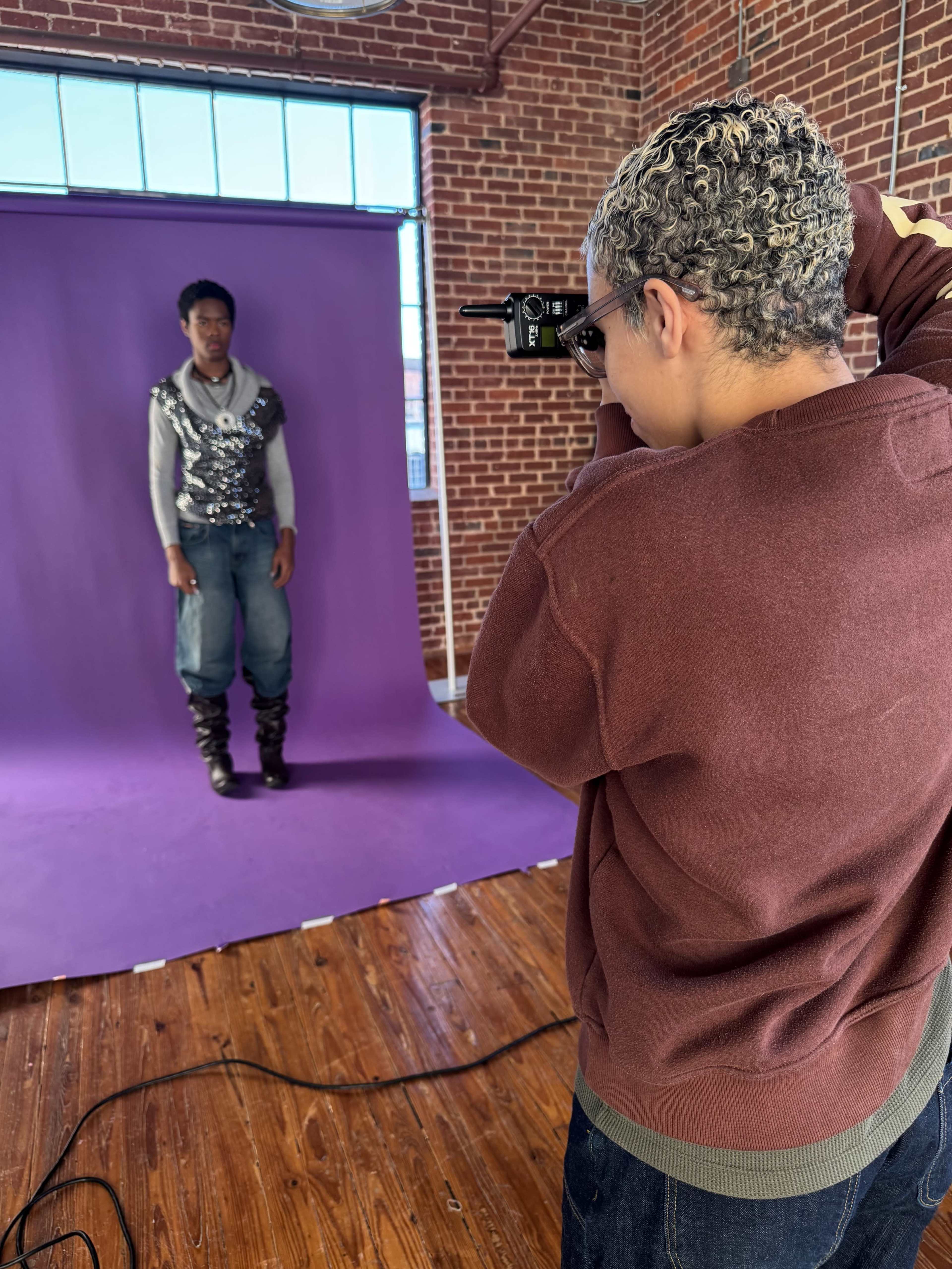 A photographer takes a picture of a model standing against a purple backdrop in a studio with exposed brick walls.
