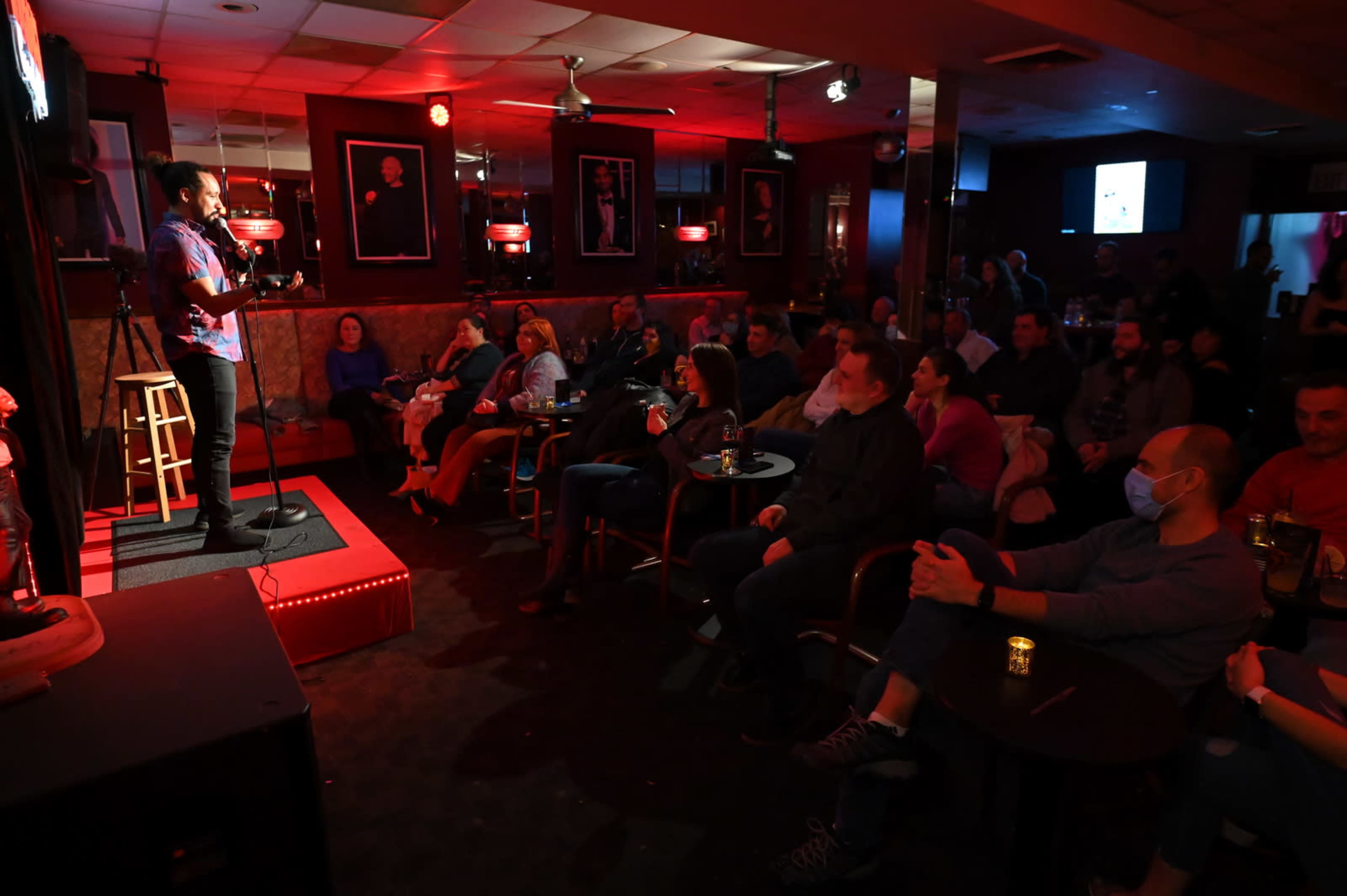 A comedian performs on stage in a dimly lit comedy club filled with an audience seated at tables, some of whom are wearing masks.