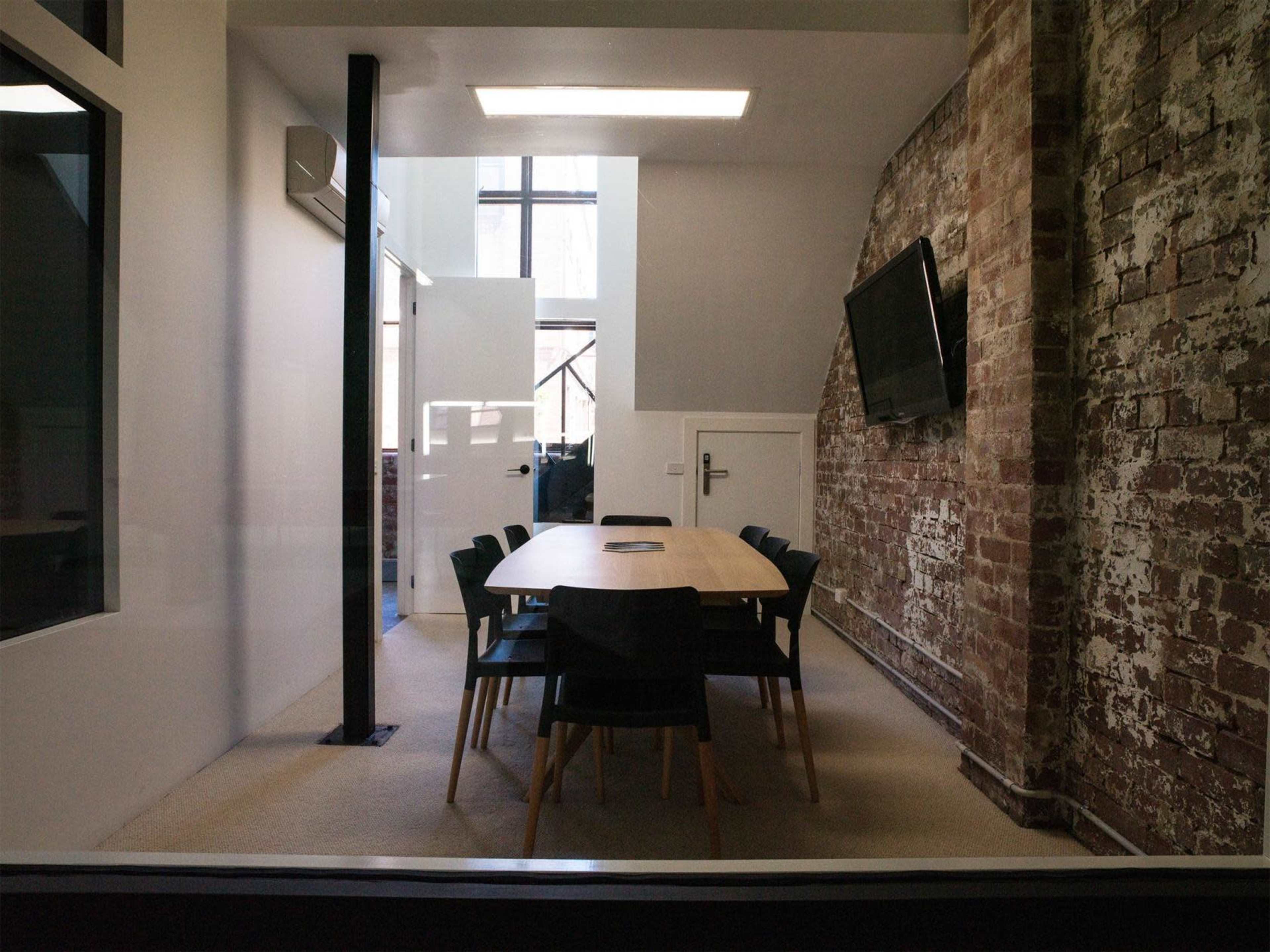 The image shows a modern conference room with a wooden table surrounded by black chairs, set against a backdrop of a brick wall and large windows.