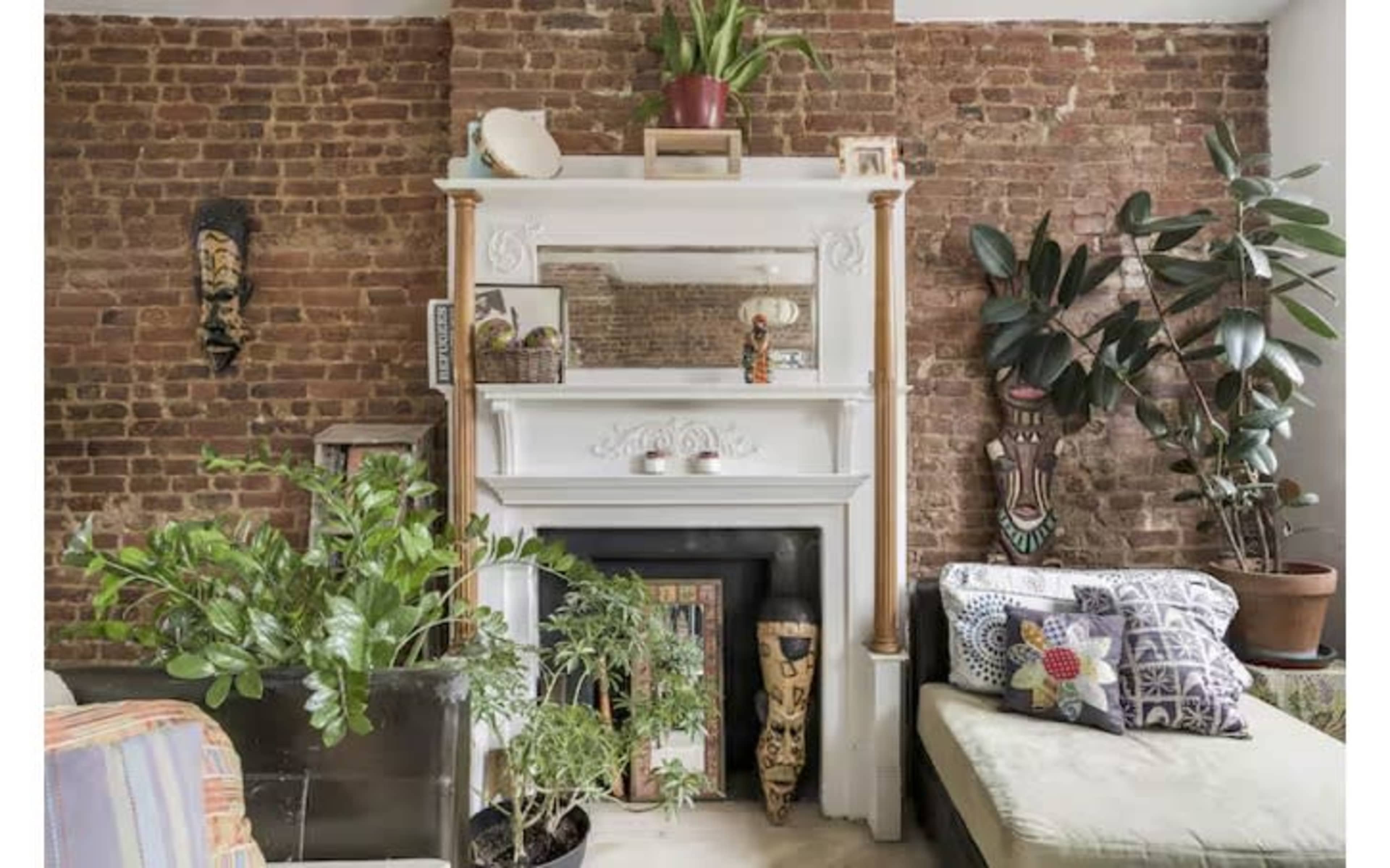 The image shows a cozy living space with a brick wall, a white fireplace mantel adorned with various decorative items, and plants in pots arranged nearby.